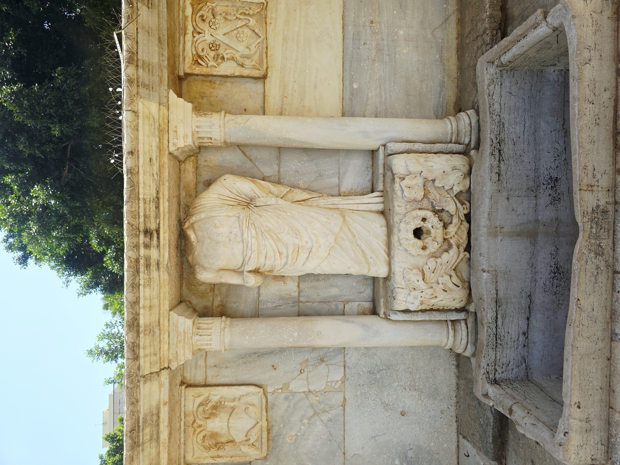 The image shows a headless marble statue of a robed figure in a niche, part of an ancient fountain structure. The statue stands in the center, flanked by fluted columns. Above and to the sides of the statue are carved marble panels with decorative motifs. Below the statue, a carved stone structure serves as a spout or decorative element, from which water would have once flowed. In front of this structure is a rectangular, stone basin or trough. The entire fountain is constructed from marble and stone, showing signs of age and weathering. The background consists of a bright sky, green trees, and a hint of a modern building behind them. This fountain is located in the Archaeological Museum of Heraklion, in the city of Heraklion, Crete, Greece.
