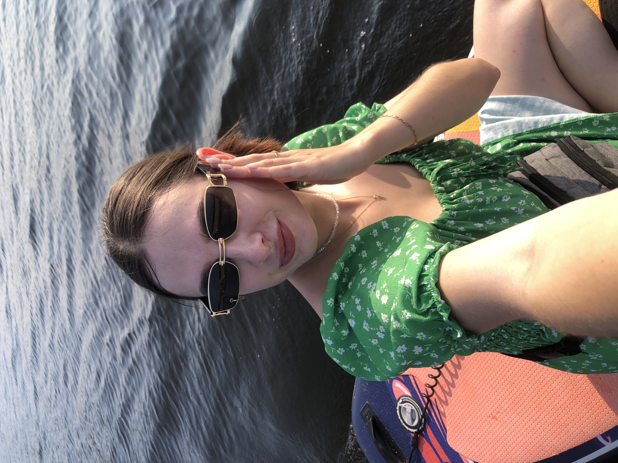 A young woman is taking a selfie while sitting on a paddleboard in a body of water. She is wearing sunglasses, a green floral dress, and a small necklace. Her right hand is touching her face. The paddleboard is orange and blue with some text and logos on it. The water is dark and rippling. The overall lighting suggests it is daytime, likely late afternoon or early evening, given the warm light. The setting appears to be a calm, open body of water, possibly a river or lake, on a clear day. Given the context of Chaikovsky, Russia, this scene is likely taking place on the Kama River.