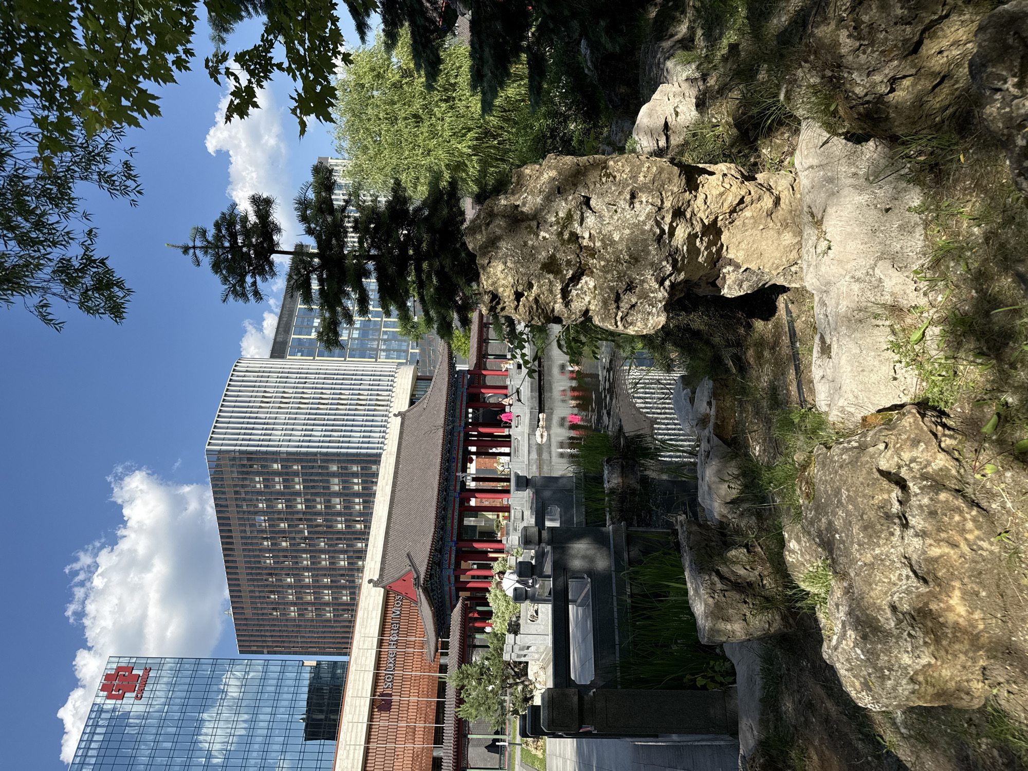 The image shows a traditional Chinese garden with a pond in the foreground, surrounded by rocks and greenery. In the background, modern high-rise buildings loom, suggesting an urban setting. A bridge crosses the pond, leading to a pagoda-style building with red columns and a dark tiled roof. Several people are visible near the bridge and on the veranda of the building, some appearing to be tourists. The sky is blue with scattered white clouds. The presence of modern architecture alongside traditional garden design indicates a contrast between old and new, likely in a city where development has integrated historical elements. Based on the architectural styles and the visible signage on the buildings, this location is the China City in Moscow, Russia.