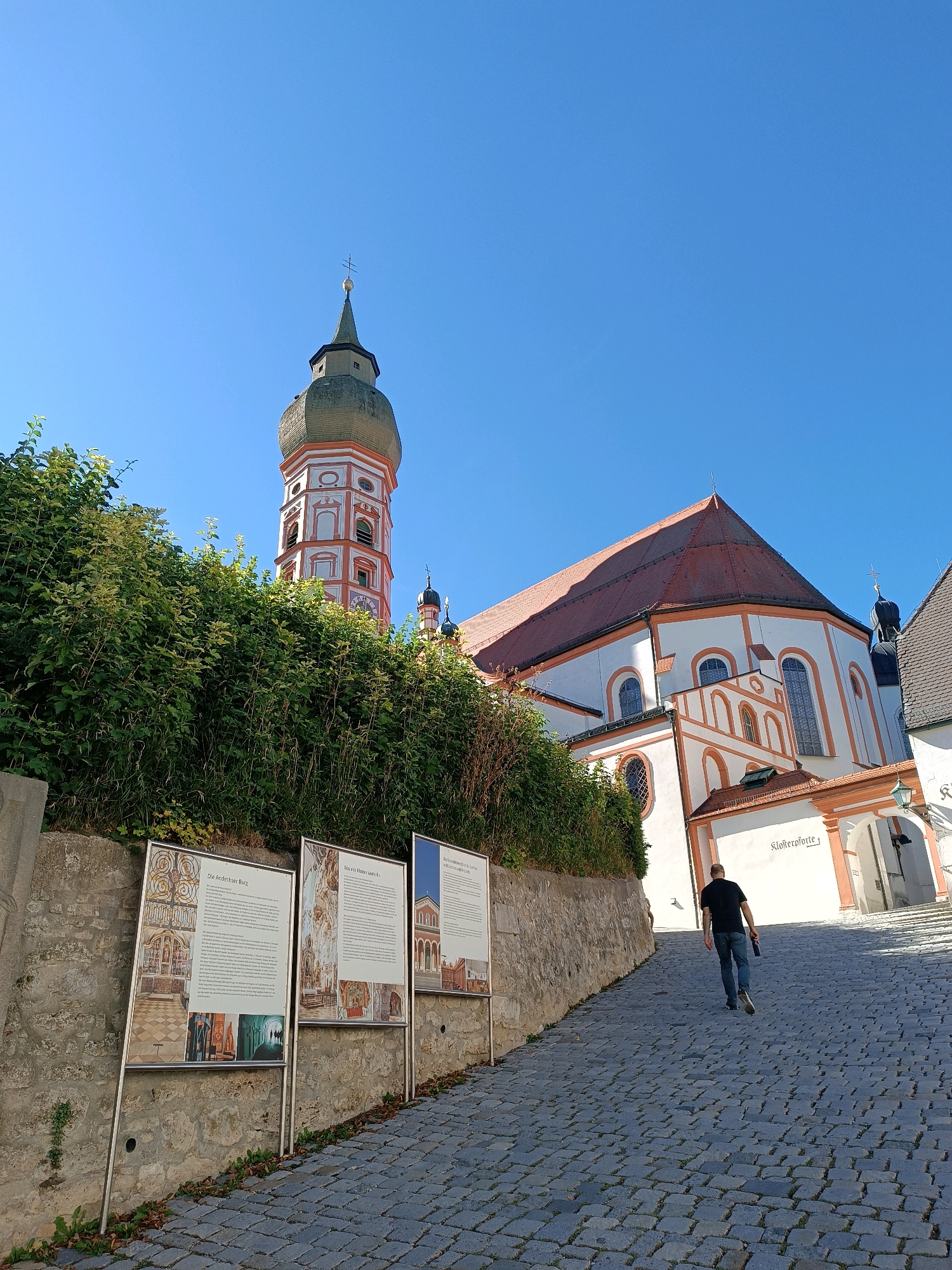 A person walks up a cobblestone path towards the Andechs Monastery in Andechs, Germany. The monastery is a large, white building with red-tiled roofs and ornate orange trim. A tall, distinctive bell tower with a green onion dome rises above the main structure. The path is lined with informational displays featuring images and text. Lush green foliage obscures part of the monastery's lower section. The sky is clear and blue.