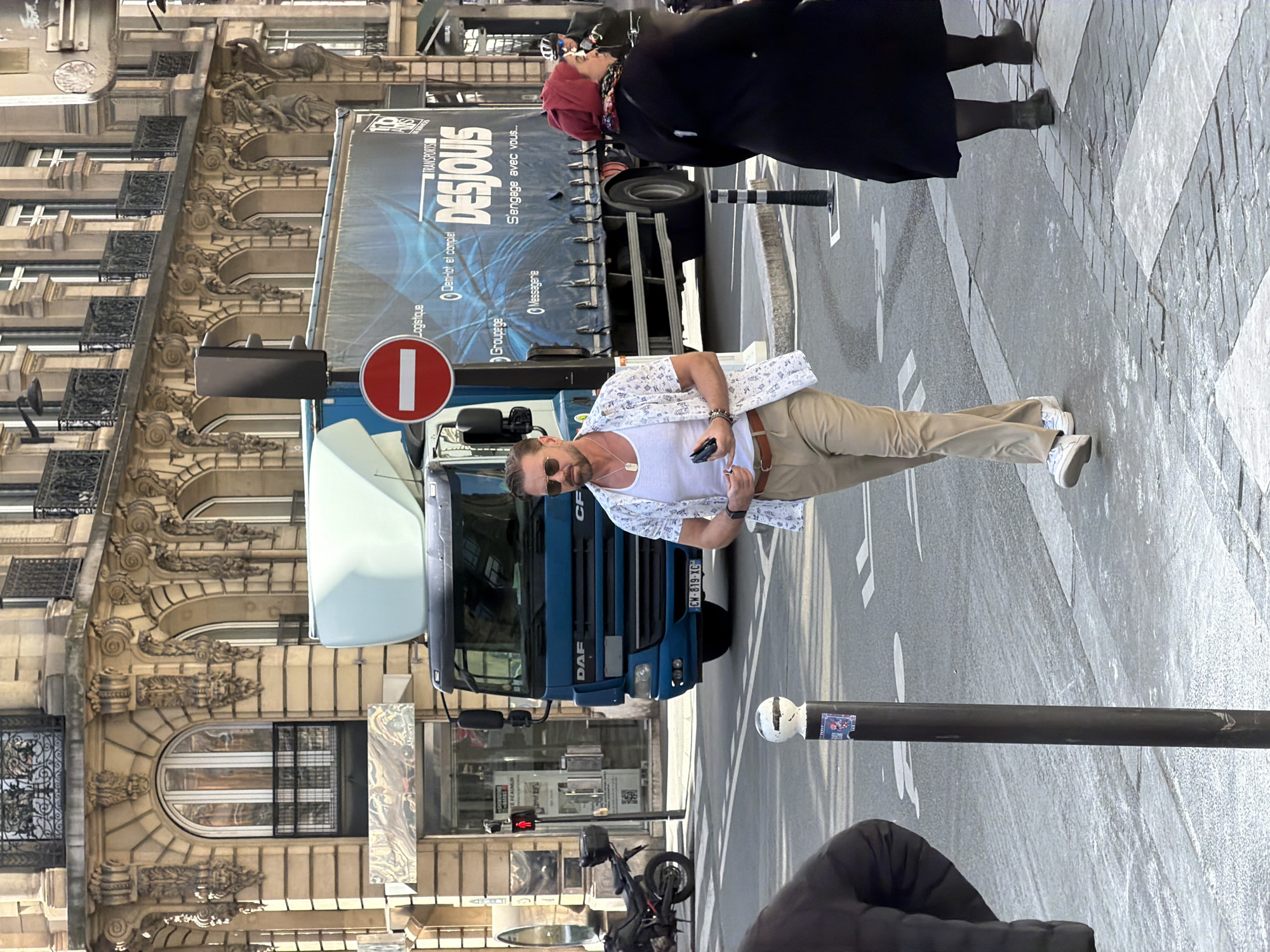 The image depicts a street scene in Paris, France. A man stands on the sidewalk near a large blue truck with the branding "TRANSPORTS DESJOUIS" prominently displayed. The truck is parked beside a building with ornate, classical architectural details. The man is wearing sunglasses, a white t-shirt, a patterned shirt, and khaki pants. He is holding a cell phone in his hand. A woman wearing a black coat is walking in the same direction as the man. There is a black pole with a white top on the right side of the image and a parked motorcycle partially visible in the foreground. A red "no entry" sign is attached to the truck. The license plate "CW-819-XC" is visible on the truck's front.