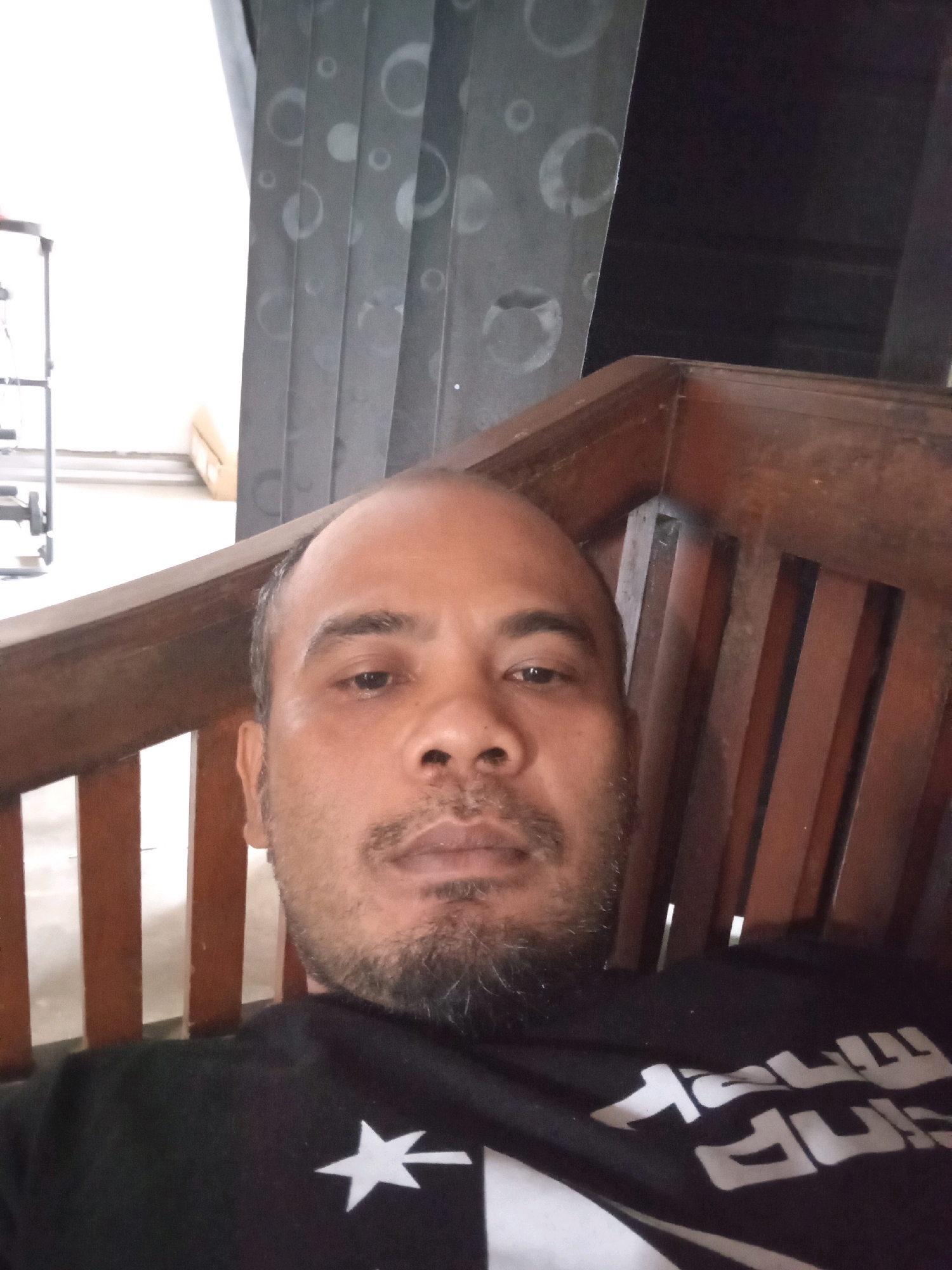 A medium shot portrait shows a bald man with a goatee and mustache sitting in a wooden chair. He is wearing a black t-shirt with a white star and writing on it. The background consists of a patterned curtain and wooden slats. The image appears to have been taken indoors, possibly in a residential setting in Kebanaran, Indonesia. The lighting is natural, coming from the left side of the frame.