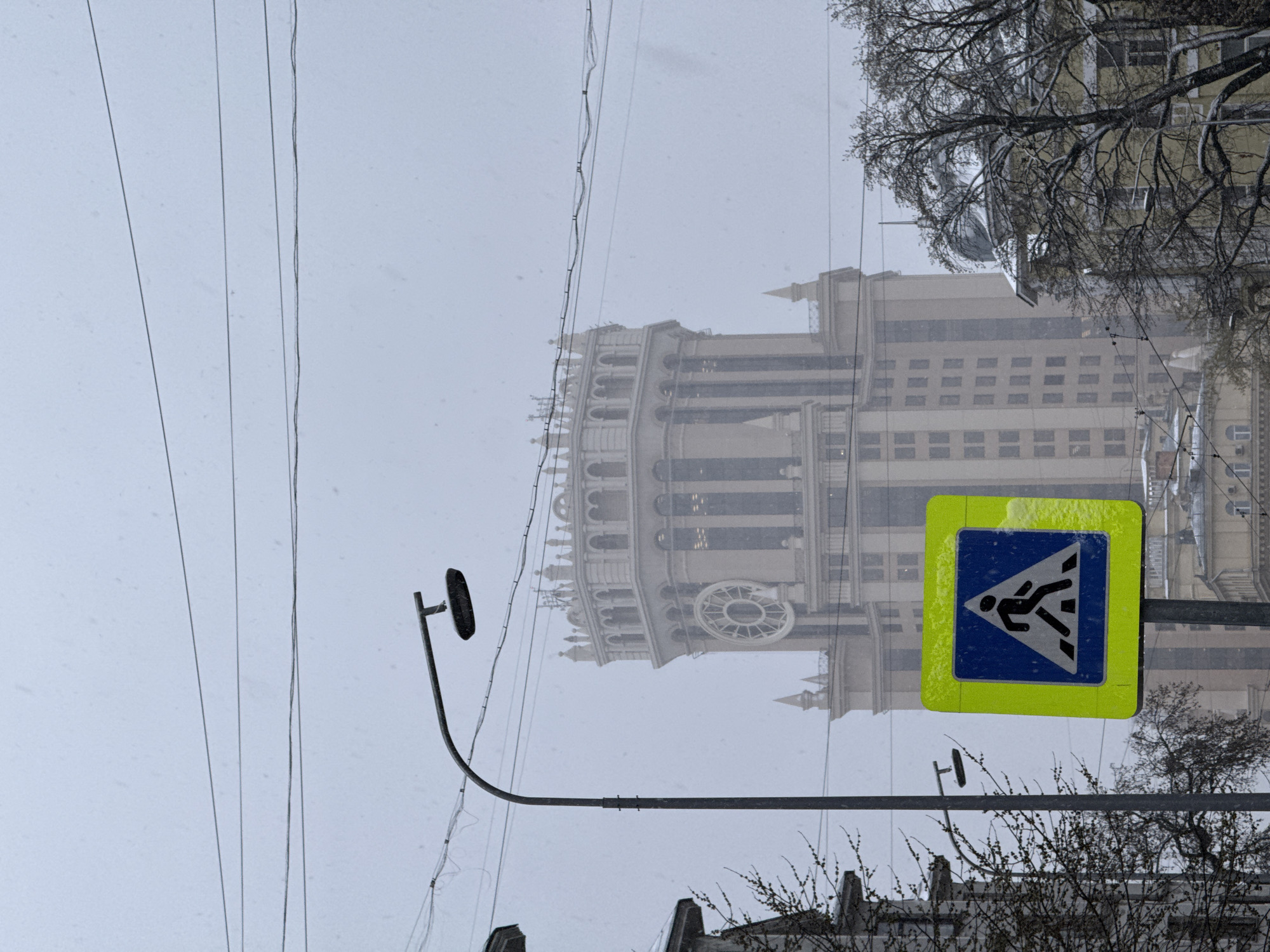 The image depicts a view of the Kudrinskaya Square Building, one of the Seven Sisters skyscrapers in Moscow, Russia, during snowy weather. A pedestrian crossing sign is visible in the foreground, featuring a blue square with a white figure walking across a crosswalk, bordered by a yellow-green square. Power lines stretch across the top left of the frame, and a street lamp is situated below the sign. Bare trees are visible on the right side of the image, with rooftops and buildings in the lower part of the frame. The Kudrinskaya Square Building is partially obscured by the snow.