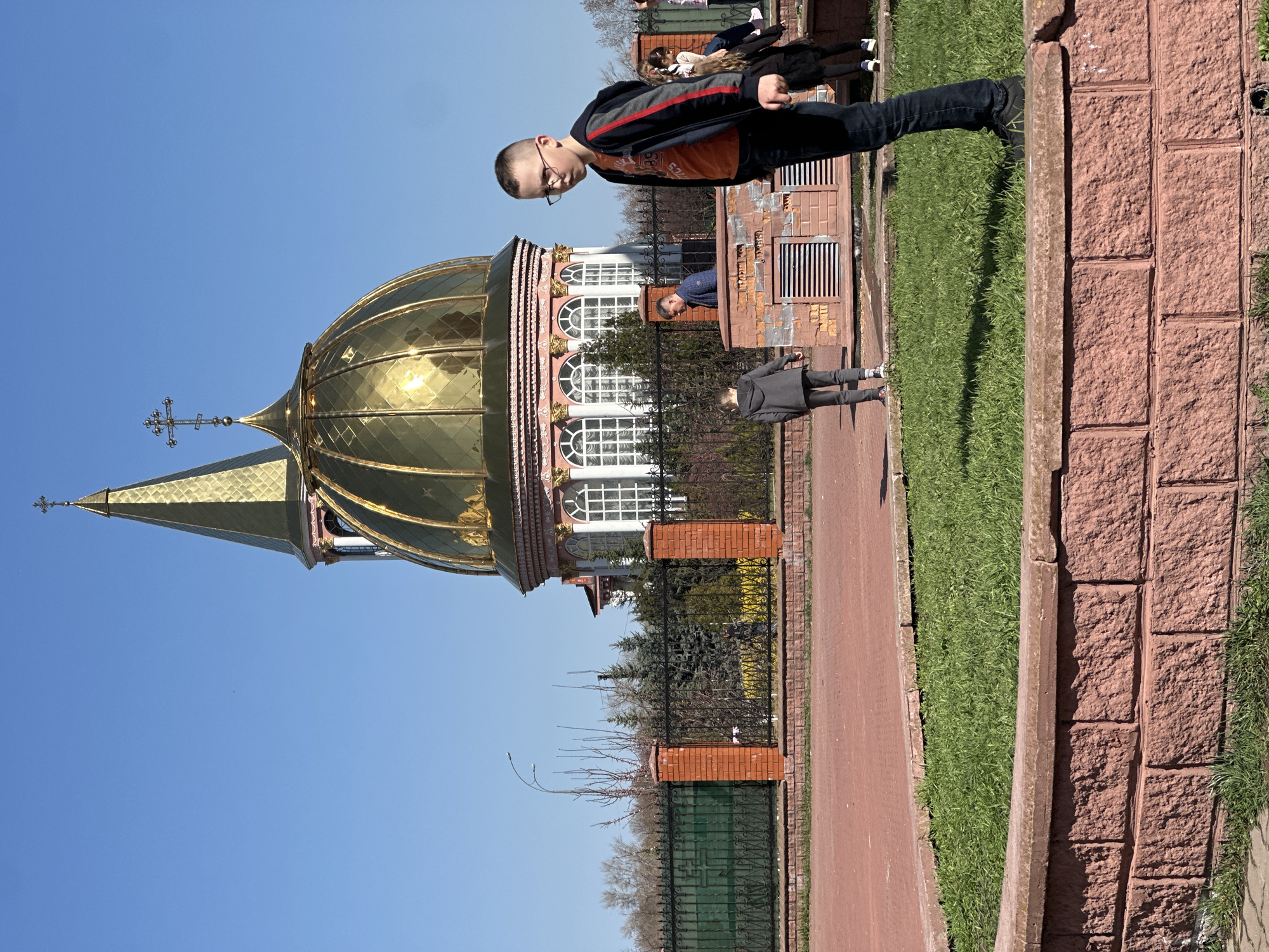 The image shows a sunny outdoor scene, likely in the city of Kyiv, Ukraine. The most prominent feature is the golden dome of a church with a cross on top. A boy with glasses, wearing a black and orange jacket and jeans, stands near the church. Other individuals are visible, including what appears to be a child in a gray coat and an adult in a blue sweater. The setting includes a red brick pathway or sidewalk, green grass, and a brick wall. There is a black metal fence with brick pillars. The church has white arched windows with pink details. A building with ventilation openings is located behind the church. The sky is clear and blue.