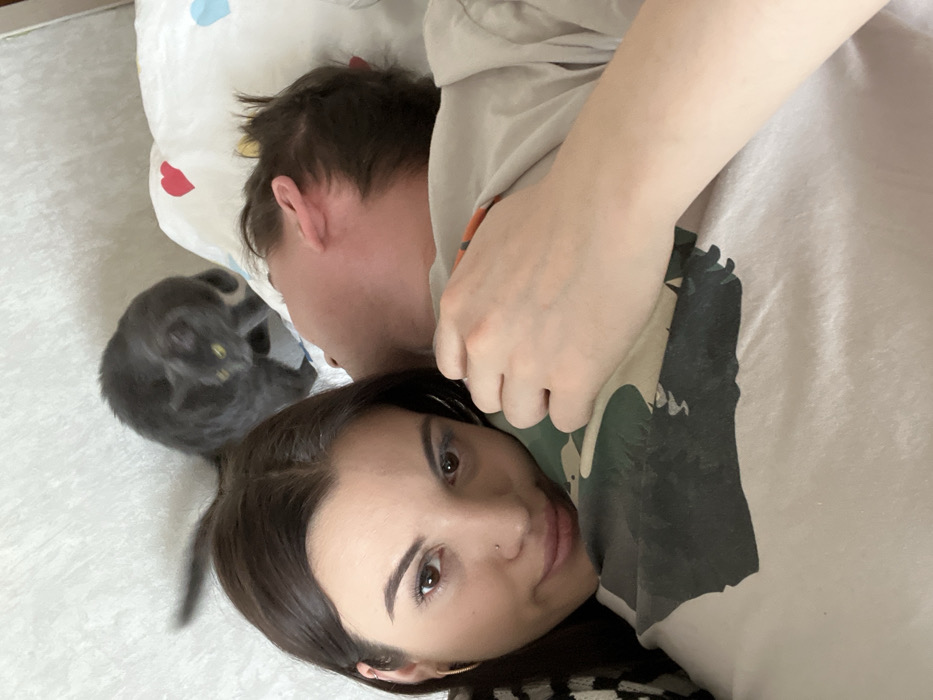 A woman with brown hair and eyes lies in bed, looking at the camera. Next to her is a man with short brown hair, facing away. A gray cat is nestled next to the man on the pillow. The woman is wearing a patterned top. The man is wearing a beige t-shirt with a print. The bed has a white pillow with small colorful patterns. The background shows a light-colored floor.
