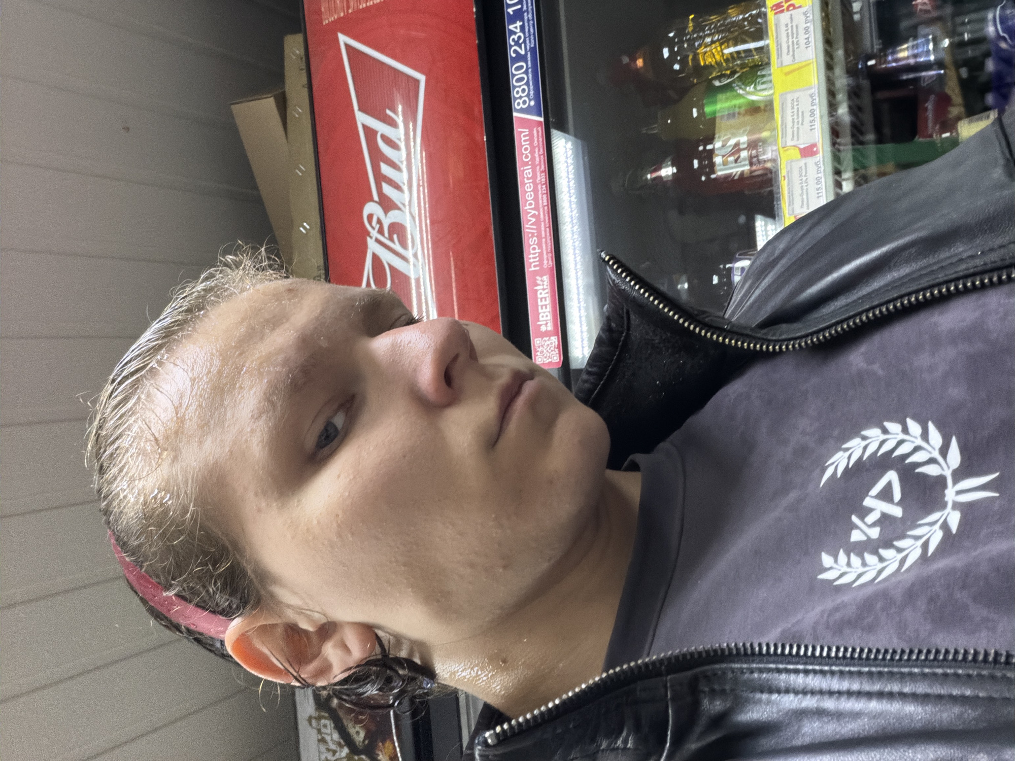A close-up, selfie-style photograph is taken from a low angle, focusing on the upper body of a person. The subject, who appears to be young, has fair skin, damp, light brown hair pulled back with a pink headband, and is sweating. They are wearing a dark grey t-shirt with a white graphic of a laurel wreath and an emblem in the center, and a black leather jacket that is zipped up. The background features a red Budweiser logo illuminated by overhead fluorescent lighting, indicating the subject is likely in a convenience store or supermarket. Below the Budweiser sign, a shelf is visible with various bottles of beverages, some with yellow price tags displaying Russian currency (руб.). A sign to the right shows a phone number: "8800 234 10..." and a website address: "https://vyberai.com/". The setting suggests an indoor environment with commercial refrigeration units. The image was captured in Chistopol, Russia.