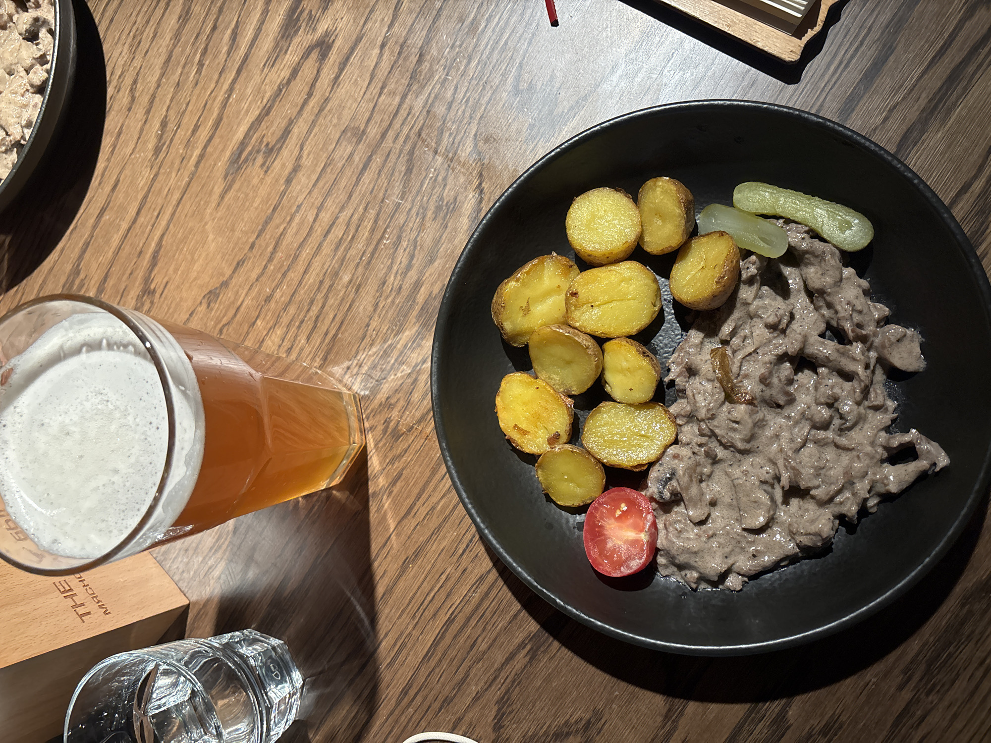 An overhead shot shows a black plate of food, a beer, and other objects on a wooden table. The plate contains fried potatoes, two pickles, a half-cherry tomato, and a portion of beef stroganoff. Next to the plate, a pint glass filled with beer has a substantial foamy head. Partially visible are a small wooden box labeled "THE MACHOIC" and an empty glass. The location is likely Moscow, Russia, given the prevalence of beef stroganoff in Russian cuisine.