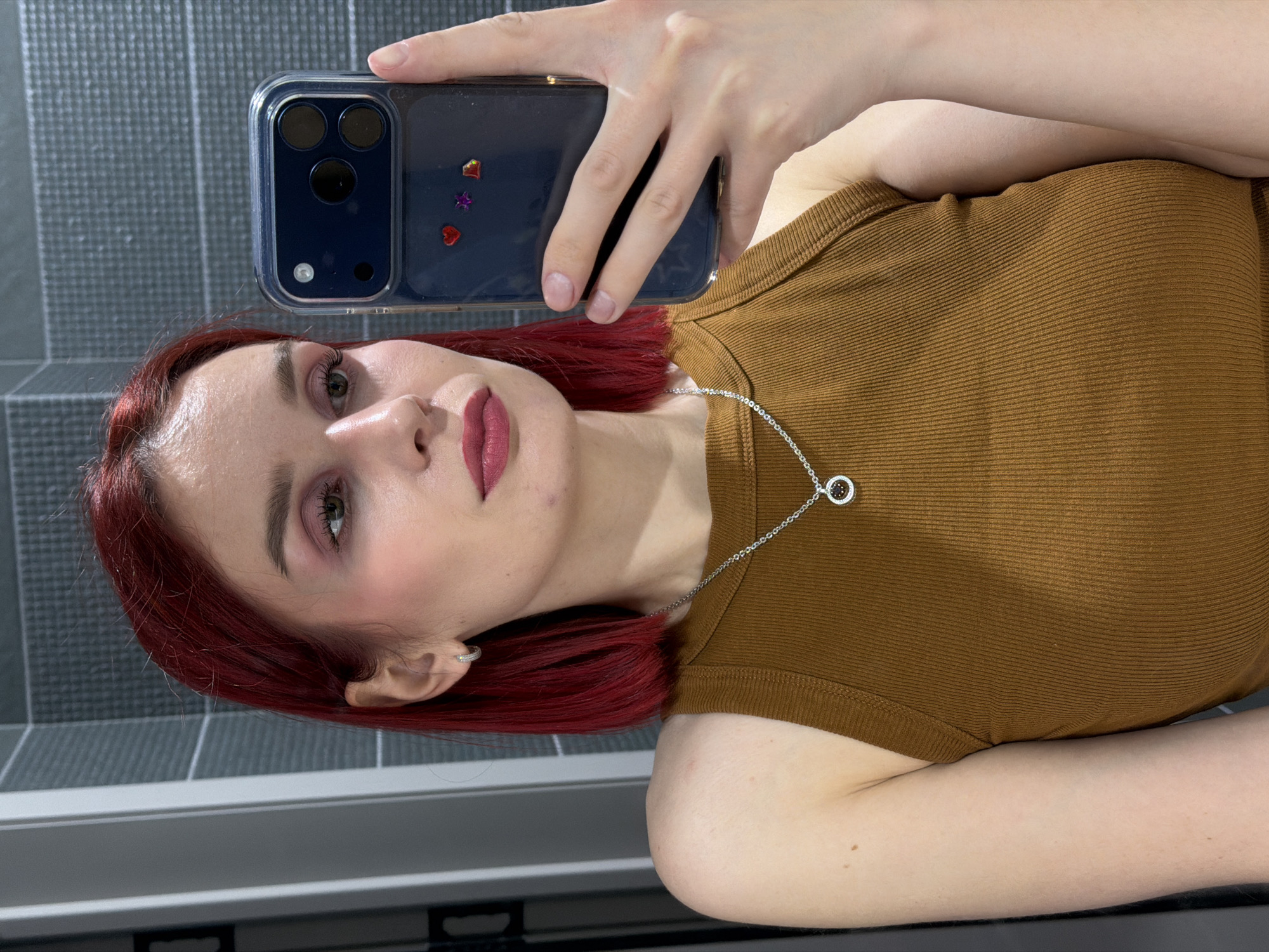 This close-up portrait-style image, likely taken in Chaikovsky, Russia, features a young woman with distinctive red/auburn hair. Her head is tilted to the right as she appears to be taking a mirror selfie or using a front-facing camera. She has fair skin, visible green eyes accentuated with pinkish eyeshadow, prominent eyebrows, and pink lipstick. A small blemish or acne is visible on her chin.
She is wearing a simple brown ribbed tank top and a silver chain necklace with a circular pendant, which contains multiple small dark or brown stones. A small silver hoop earring is visible in her left ear.
The woman is holding a smartphone, identifiable by its triple camera array, within a clear case. The case is adorned with small, shiny heart and star-shaped embellishments in shades of red, purple, and orange. Her hand and fingers are visible gripping the device.
The background consists of a dark gray tiled wall with lighter grout lines arranged in a grid pattern, suggesting a modern interior space, potentially a bathroom. Below the tiled wall, a light-colored horizontal surface is visible. The lighting is even, typical of an indoor setting. The woman looks towards the camera with a neutral expression. There is no visible text in the image.