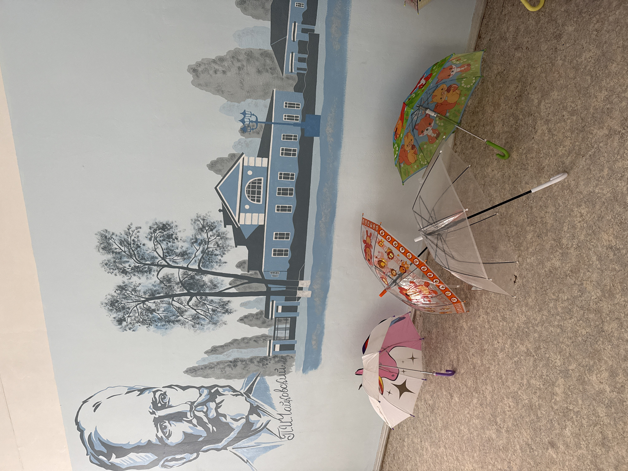The image shows a corner of a room with a painted mural on the wall and several open umbrellas resting on the floor. The mural depicts a landscape with a large building, trees, and clouds, all rendered in shades of blue and gray. At the bottom left corner of the mural is a portrait of Pyotr Ilyich Tchaikovsky. In Votkinsk, Russia, Tchaikovsky's birthplace features a museum complex, suggesting this might be a related site. The umbrellas, each with different designs (clear, cartoon, and unicorn-themed), are leaning against the wall and lying on the floor in the corner where the mural ends and the floor begins. The floor is covered with a speckled, neutral-colored linoleum or similar material. The scene appears to be indoors, potentially in a hallway or room with a cultural or educational theme related to Tchaikovsky. There are no people visible. The scene suggests an area where umbrellas might be left to dry, especially after rainy weather.