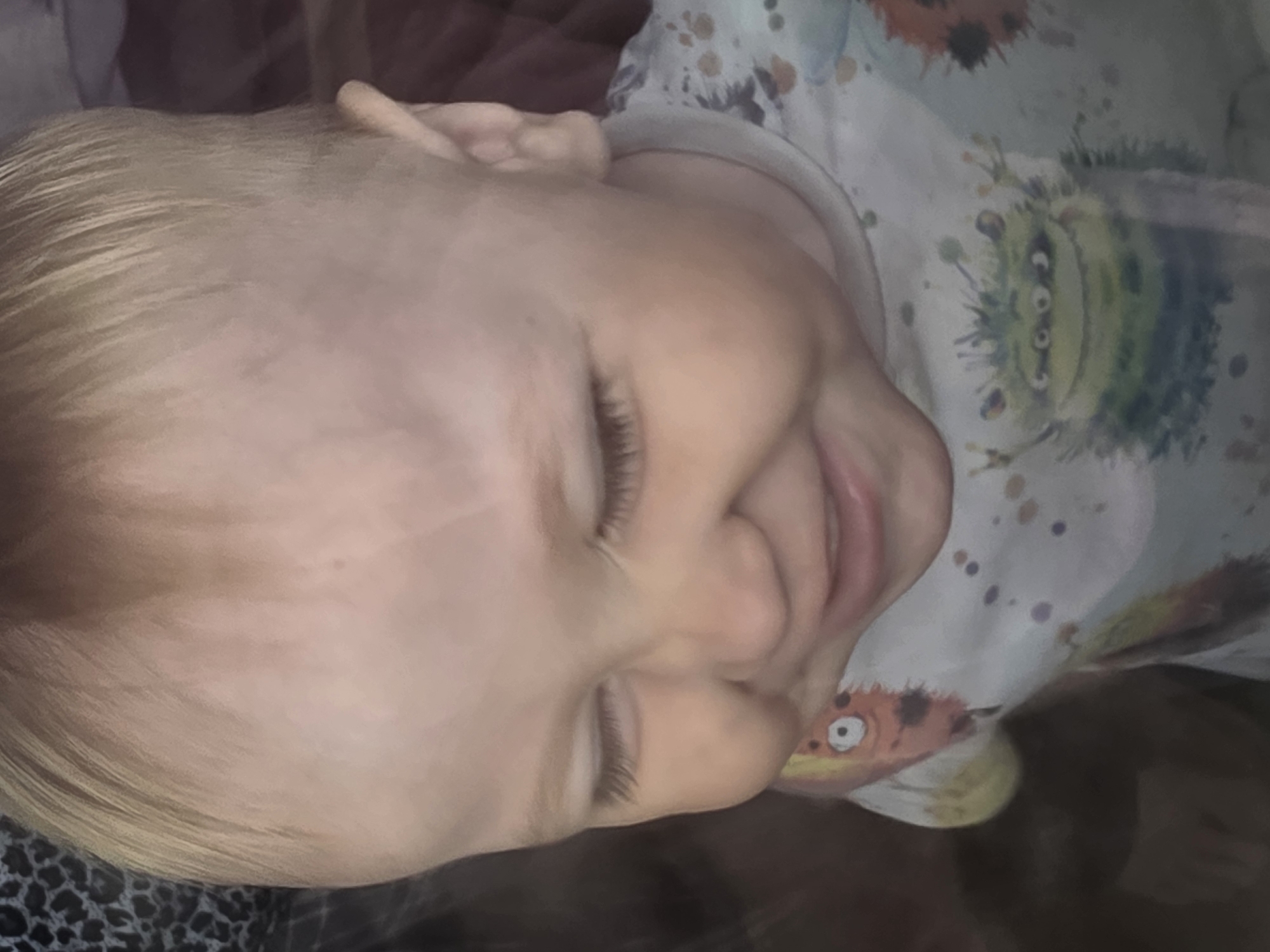 A fair-skinned child with blonde hair is lying down, eyes closed. The child is wearing a light-colored shirt with cartoon monsters printed on it. The location is unknown.