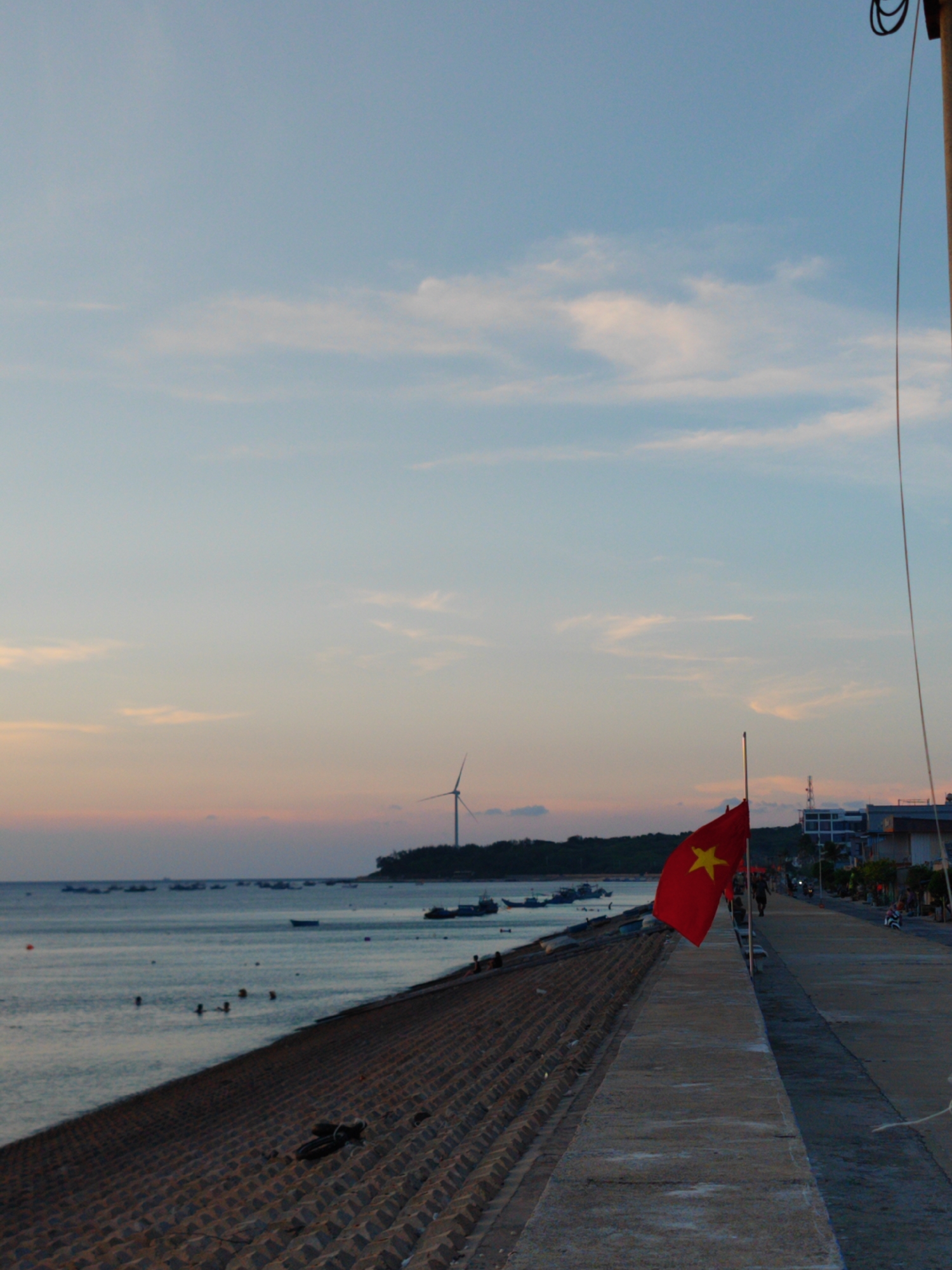 Captured in the Phú Quý Special Administrative Region, Vietnam, the photograph depicts a waterfront scene at dusk. A red flag emblazoned with a gold star, the flag of Vietnam, is prominently displayed on the right. The flag sits adjacent to a concrete walkway or promenade. To the left of the walkway, a concrete sea defense structure slopes down to meet the water. In the distance, numerous boats dot the sea, with a landmass visible, marked by a modern wind turbine. People are visible along the walkway and in the water. The sky is a gradient of pale blues and soft orange hues, suggesting either sunrise or sunset.