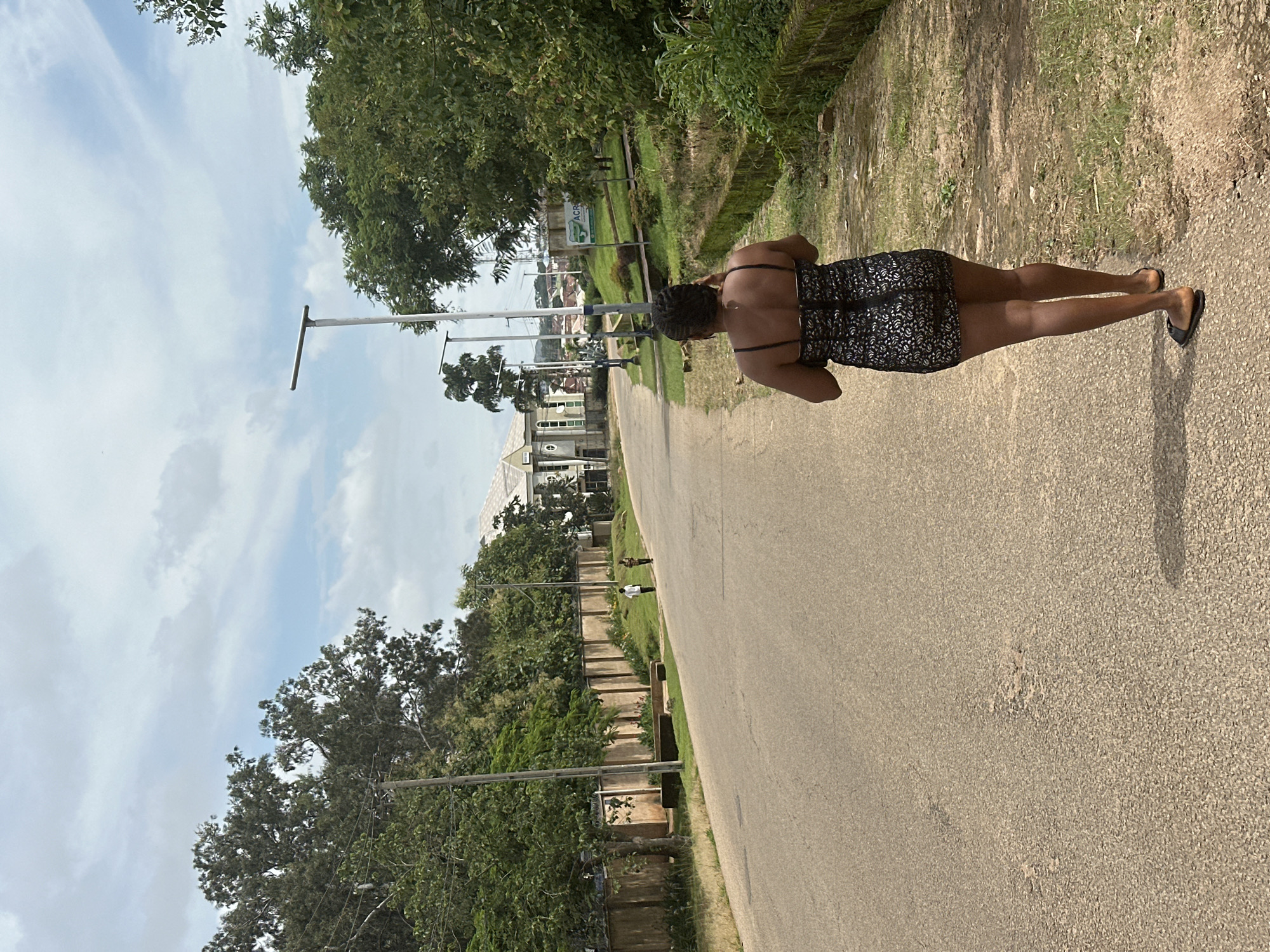A woman with dark skin and braided hair, wearing a short black and white patterned dress and flat black sandals, walks away from the camera on a paved road in Jos, Nigeria. The road is lined with green trees and grass. A wall with a barbed wire fence runs along the left side of the road. In the distance, a large building with a light-colored facade and a prominent dome is visible. The sky is cloudy.