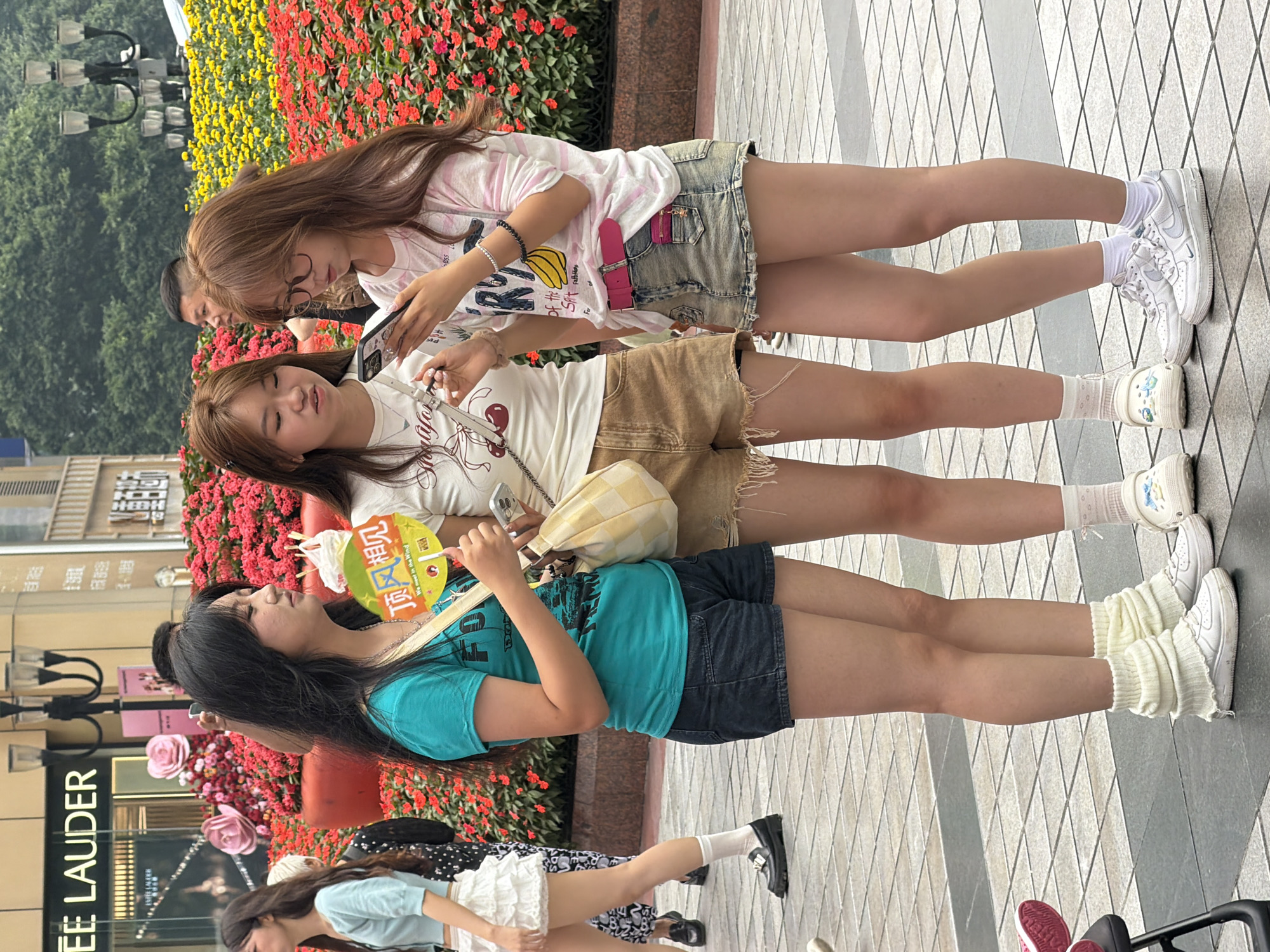 Three young women are standing on a tiled pedestrian walkway in front of a flower display in Yuzhong District, China. The woman on the left wears a turquoise t-shirt and black shorts, holding a yellow fan with Chinese characters. The woman in the middle has on a white t-shirt and brown shorts, looking at her phone. The woman on the right wears a pink and white striped t-shirt and a denim skirt, also looking at her phone. Behind them is a vibrant display of red and yellow flowers. In the background, a street lamp and the facade of a building with "LEE LAUDER" visible are present. The overall environment suggests an urban outdoor setting, possibly a commercial area.