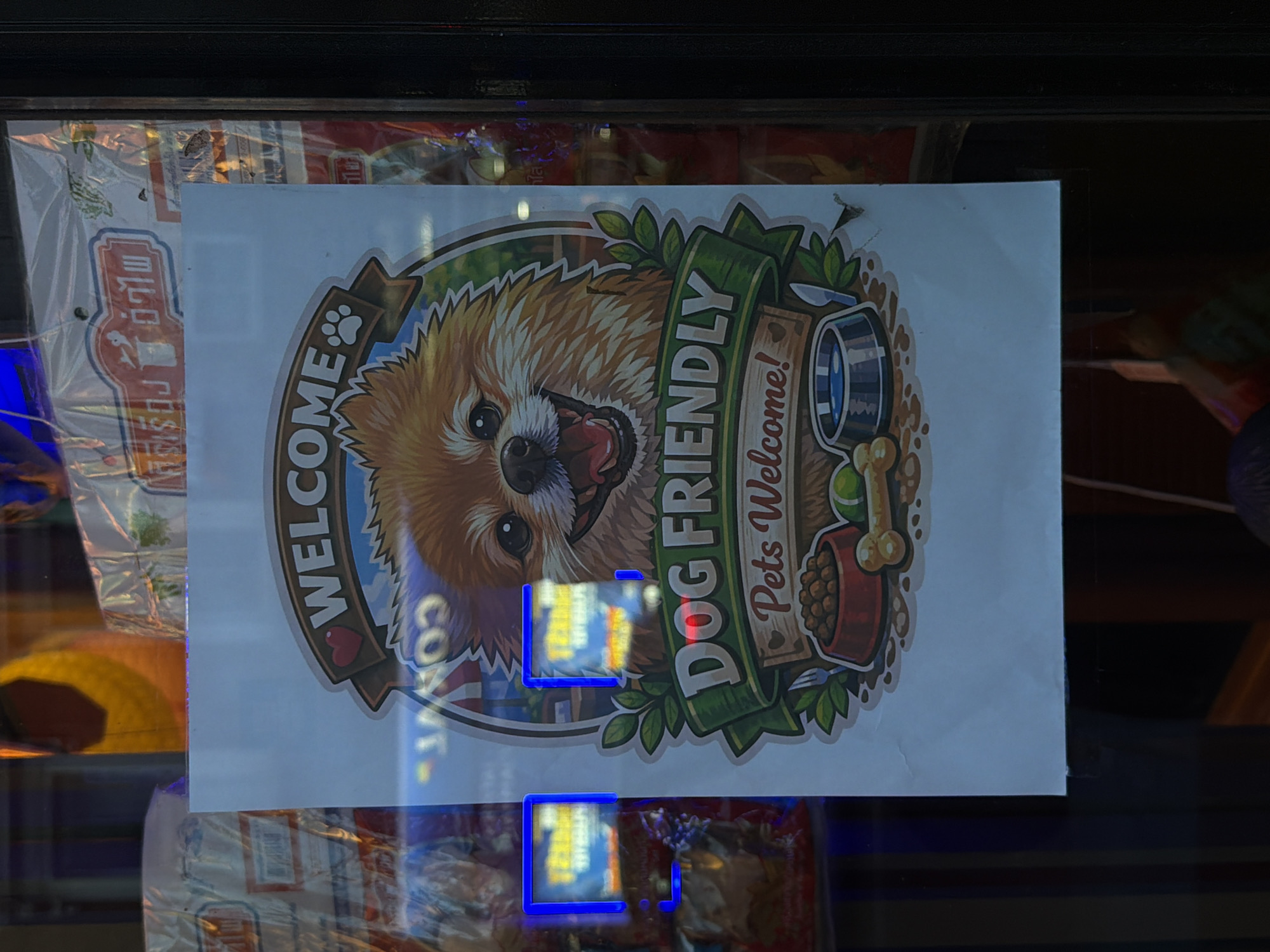 The image depicts a "Dog Friendly" sign affixed to what appears to be a glass window or door. The sign features an illustration of a Pomeranian dog along with the text "WELCOME" and "DOG FRIENDLY - Pets Welcome!" There are images of a dog bowl, bone, and dog food bowl. The sign is surrounded by what seems to be commercial packaging, potentially related to pet supplies or food. It is difficult to deduce the exact location in London, UK, without further contextual clues or identifiers visible in the broader scene.