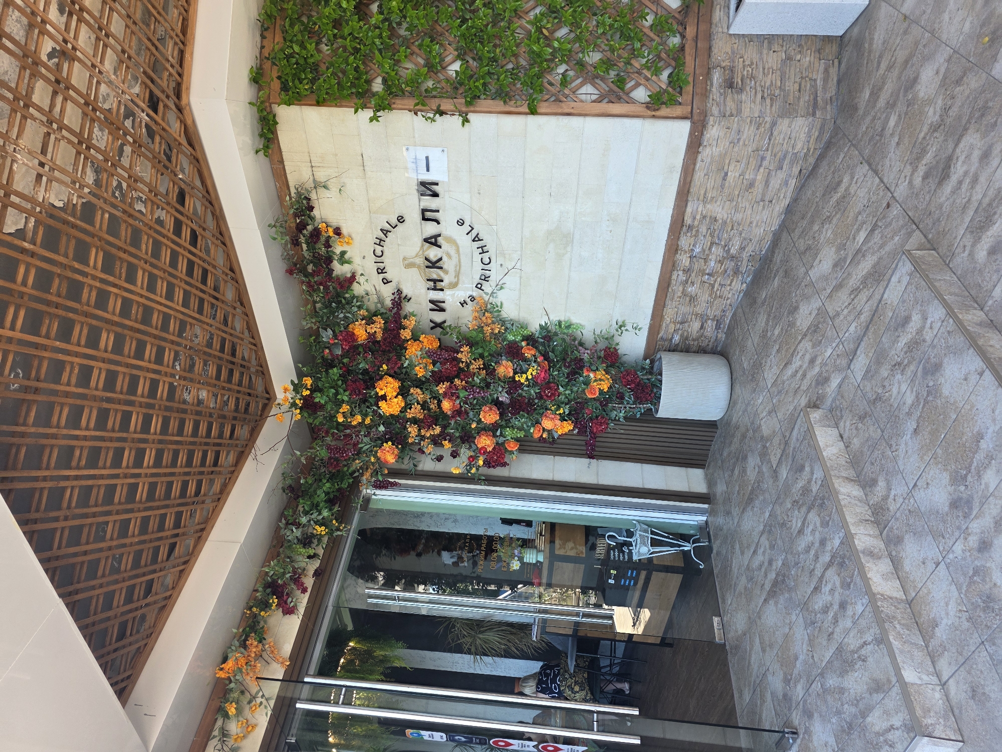 The image depicts the exterior entrance of "Хинкали на Причале" (Khinkali on the Pier), a restaurant located in Sochi, Russia. The building features light-colored, horizontal stone block walls, with a lower section clad in stacked, rough-hewn stone tiles. Above the entrance, a white soffit transitions to a ceiling partially covered by a decorative grid of brown wooden slats, revealing a darker, uneven surface underneath.
An extensive artificial floral and greenery arrangement dominates the facade. It features dense clusters of orange and deep red flowers, intermingled with green foliage and dark, berry-like clusters (resembling grapes). This arrangement originates from a large, light-colored cylindrical planter on the ground to the right of the entrance and sprawls upwards and across the wall, encompassing a significant portion of the facade. To the far right, a wooden lattice is covered with green climbing vines.
The restaurant's name, "на PRICHALE," is inscribed in black capital letters above a black, stylized khinkali dumpling icon, followed by "ХИНКАЛИ-" below it, and "на PRICHALE" again in a semi-circular arrangement beneath the icon, all mounted on the light stone wall. A small, blank rectangular sign is affixed next to "ХИНКАЛИ-".
The entrance consists of modern double glass doors. Through the left glass panel, multiple stickers, possibly indicating services or payment methods, are visible. The interior is partially visible through the glass, showing a person with blonde hair seated at a high-top table, facing away from the camera. Interior decor includes potted plants and a white, ornate metal stand near the entrance, likely for umbrellas. A sign inside, visible through the glass, indicates "РЕЖИМ РАБОТЫ" (Operating Hours) as "08.00 - 00.00" (8 AM to 12 AM) "ЕЖЕДНЕВНО" (Daily).
The ground in front of the entrance is paved with beige and brown mottled square tiles, leading to a single step with a metal trim at the edge before the building's threshold. A rectangular white planter is situated on the far right of the paved area. The scene is illuminated by natural daylight.