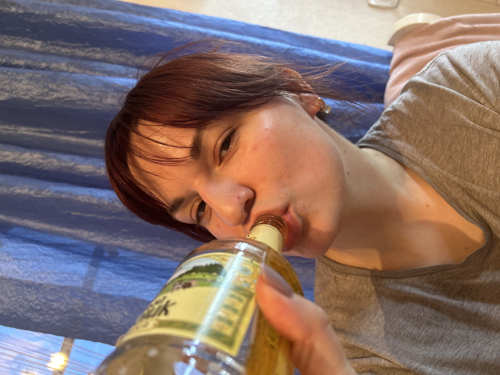 The image presents a close-up, angled portrait of a person drinking directly from a glass bottle. The subject, likely female, has short, reddish-brown hair and fair skin. They are wearing a casual grey t-shirt. Their gaze is directed towards the viewer, with a relaxed, direct expression as they sip from the bottle. A small earring is visible in their right ear and a ring is on their finger.
The person is actively drinking from a clear glass bottle, holding it with their right hand. The bottle contains a light yellow or amber-colored liquid. Its label is light-colored, featuring a landscape illustration with trees and hills. Partially visible text on the label includes Cyrillic letters "Вин" (suggesting "Вино," meaning wine) and "ЙК," indicating an alcoholic beverage produced in Russia, consistent with the provided location context of Фоки, Russia.
The background is dominated by a textured, wavy blue fabric, possibly a curtain or blind, running horizontally. Above this blue element, a lighter, indistinct surface is visible. To the upper right, a soft pink or light brown fabric, perhaps a blanket or pillow, is partially seen. Below the blue fabric, natural light suggests the presence of a window. The overall lighting is bright and even, indicative of daytime. The composition is dynamic, with the subject positioned diagonally across the frame, conveying a candid moment.