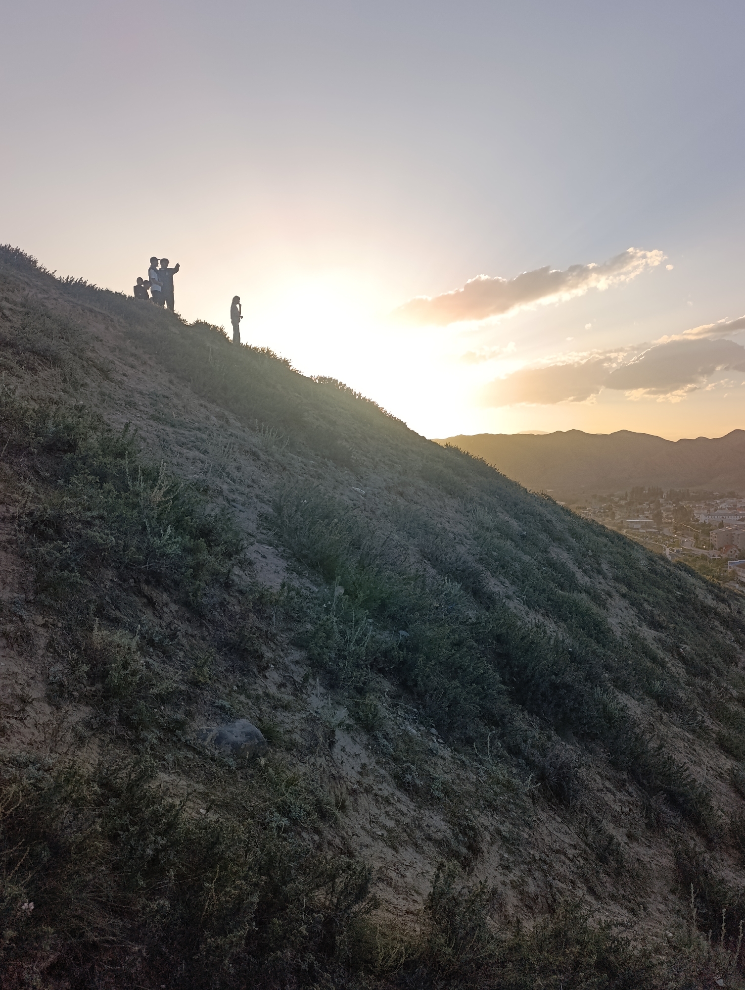 A group of people stand silhouetted on a steep, grassy hillside overlooking a city nestled in a valley at sunset. The sun, low on the horizon, casts a warm glow across the sky, illuminating wispy clouds and the distant, hazy outlines of mountains. The hillside itself is covered in sparse green vegetation and dry, sandy soil, with a few larger rocks visible.
Four figures are grouped together on the upper part of the slope. Two appear to be standing close, one with an arm raised, possibly pointing towards the view or gesturing. Another figure stands slightly apart to the right, also in silhouette. The overall mood is peaceful and contemplative, with the individuals seemingly enjoying the panoramic vista as the day draws to a close. The light suggests it is either early morning or late evening, with the warm tones of the sky leaning towards sunset. The presence of a city and mountainous terrain provides context for the location, which is Naryn City, Kyrgyzstan. No text is visible in the image.