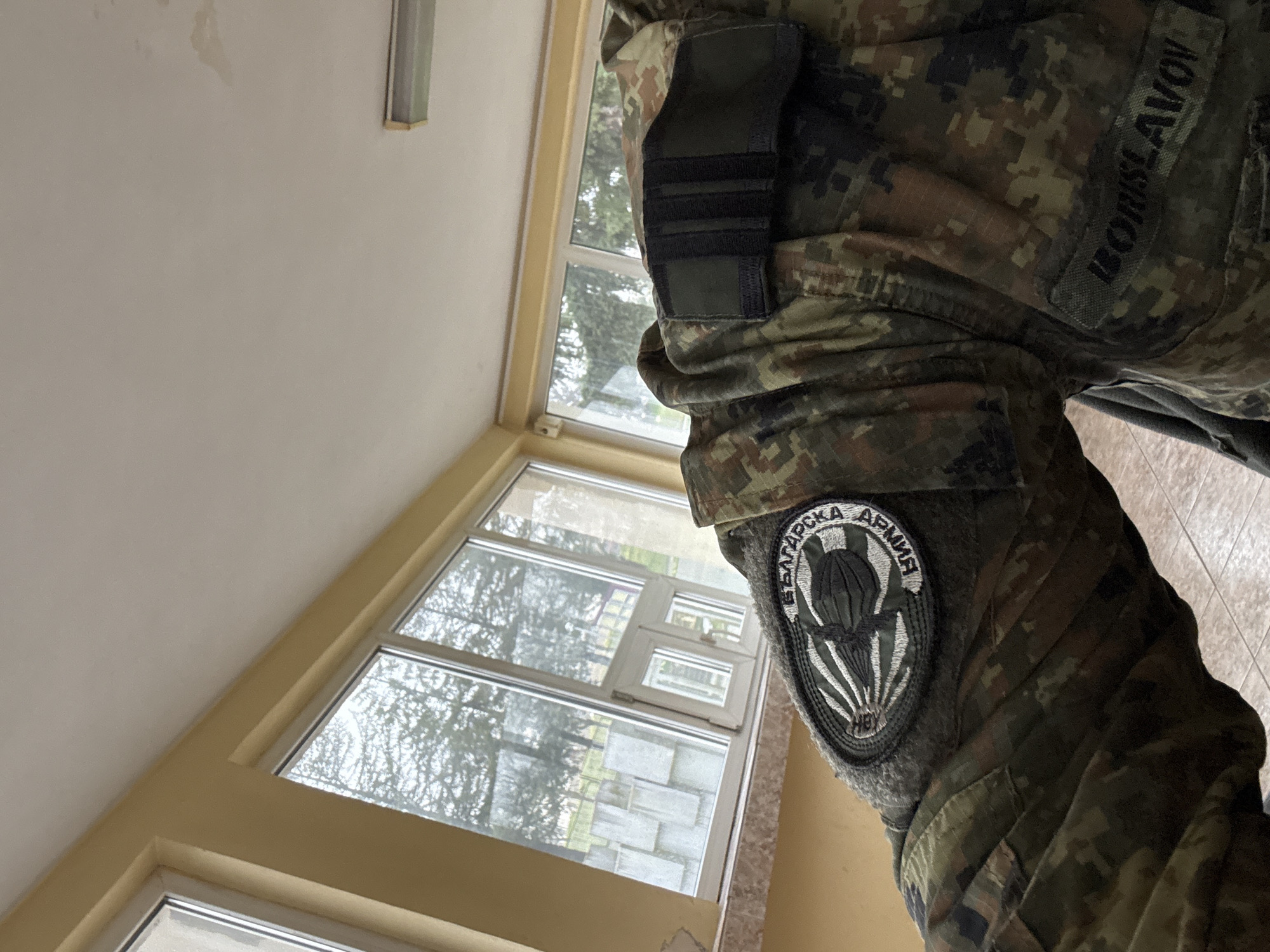 The image shows the right arm and shoulder of a person wearing a camouflage military uniform. On the sleeve is a patch with the words "БЪЛГАРСКА АРМИЯ" (Bulgarian Army) surrounding a parachute emblem. A name tag reading "BORISLAVOV" is visible above the pocket on the upper right chest area. The person is indoors, standing near a window. The window offers a view of an outside area with trees and what appears to be some kind of structure, possibly a building or monument. Based on the architecture and the "Bulgarian Army" patch, the location is likely in Bulgaria. The image does not provide enough information to precisely pinpoint Veliko Tarnovo as the specific location.