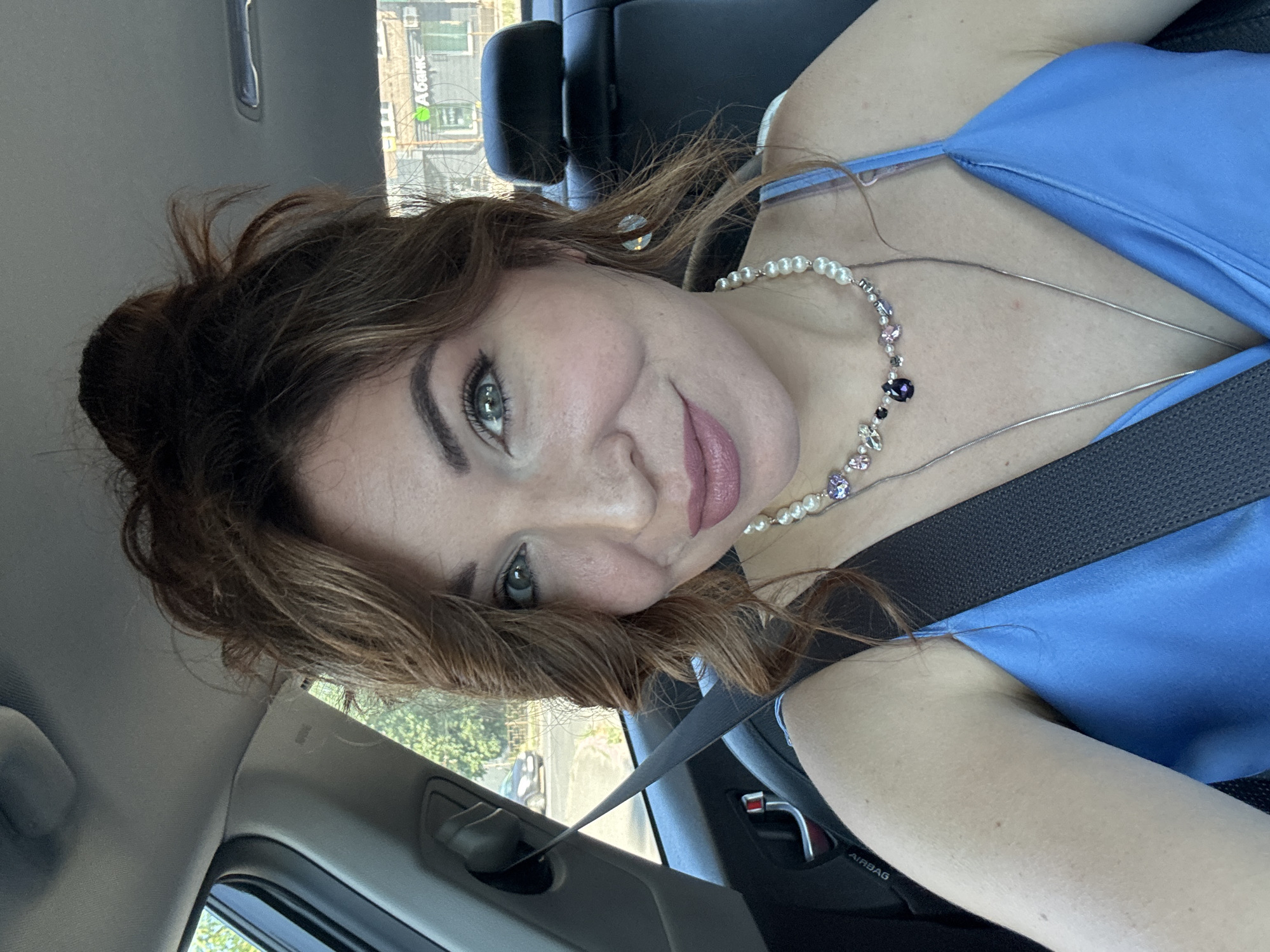 A woman is inside a car, in the driver's seat, and is taking a selfie. She has brown hair styled in a messy bun, green eyes, and is wearing a blue dress and a pearl necklace. The background shows a car interior with a seatbelt across her chest and a glimpse of trees and buildings outside the window, suggesting an urban environment. The building in the background has a green sign with text in Cyrillic script, "А-Банк", which is a bank in Ukraine. The image was taken in Kryvyi Rih, Ukraine.