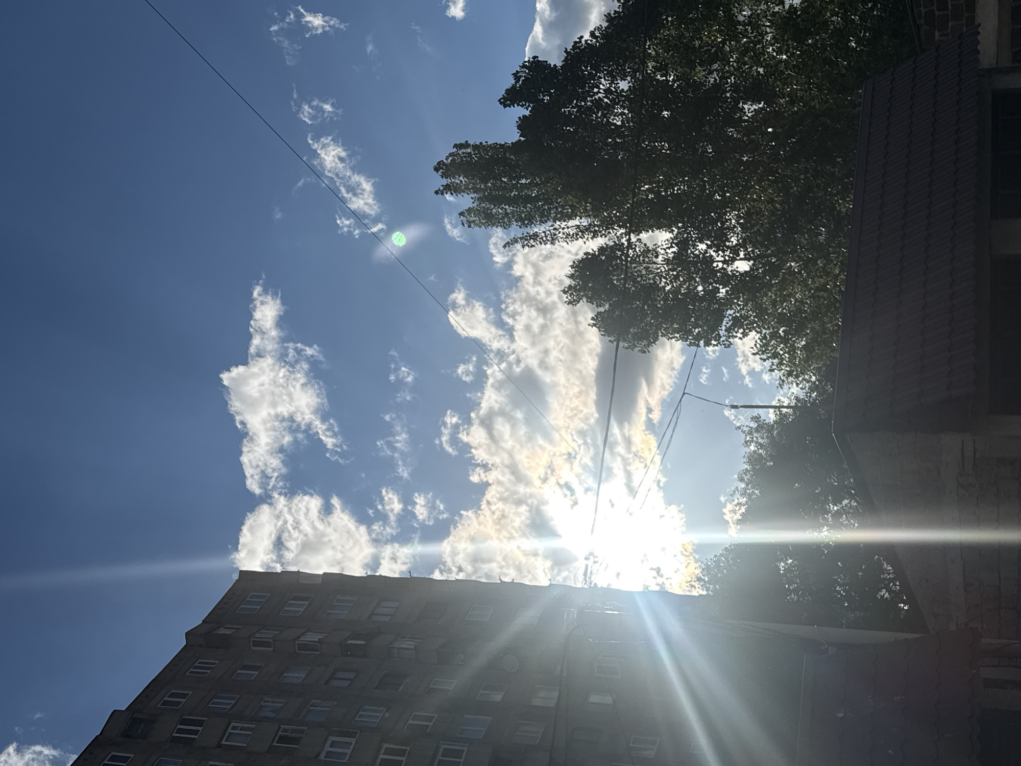 The image captures a vibrant, sun-drenched urban or suburban scene from a low-angle perspective, looking upwards towards the sky in Abovyan, Armenia. The dominant element is the bright blue sky, peppered with fluffy white clouds. The sun is intensely bright, positioned in the top-right quadrant of the frame, creating significant lens flare that radiates across much of the image and causes parts of the foreground elements to appear silhouetted. A distinct green circular lens artifact is visible in the sky near the top-left.
In the bottom-left portion of the frame, a multi-story building, likely an apartment or office block, is visible. It is constructed from a grey, possibly concrete, material and features numerous rectangular windows, some appearing open or reflecting the bright light. Towards the bottom-right, a section of another structure with a dark, ridged tiled roof and brickwork is present. Dark green foliage from trees can be seen along the top edge and extending down the right side, partially obscuring the sky and contributing to the silhouetted effect. Thin, dark utility wires traverse the sky diagonally.
The scene suggests a clear and sunny day, likely during the late morning or early afternoon given the high sun position. There are no people or specific activities depicted. No discernible text is visible within the image.