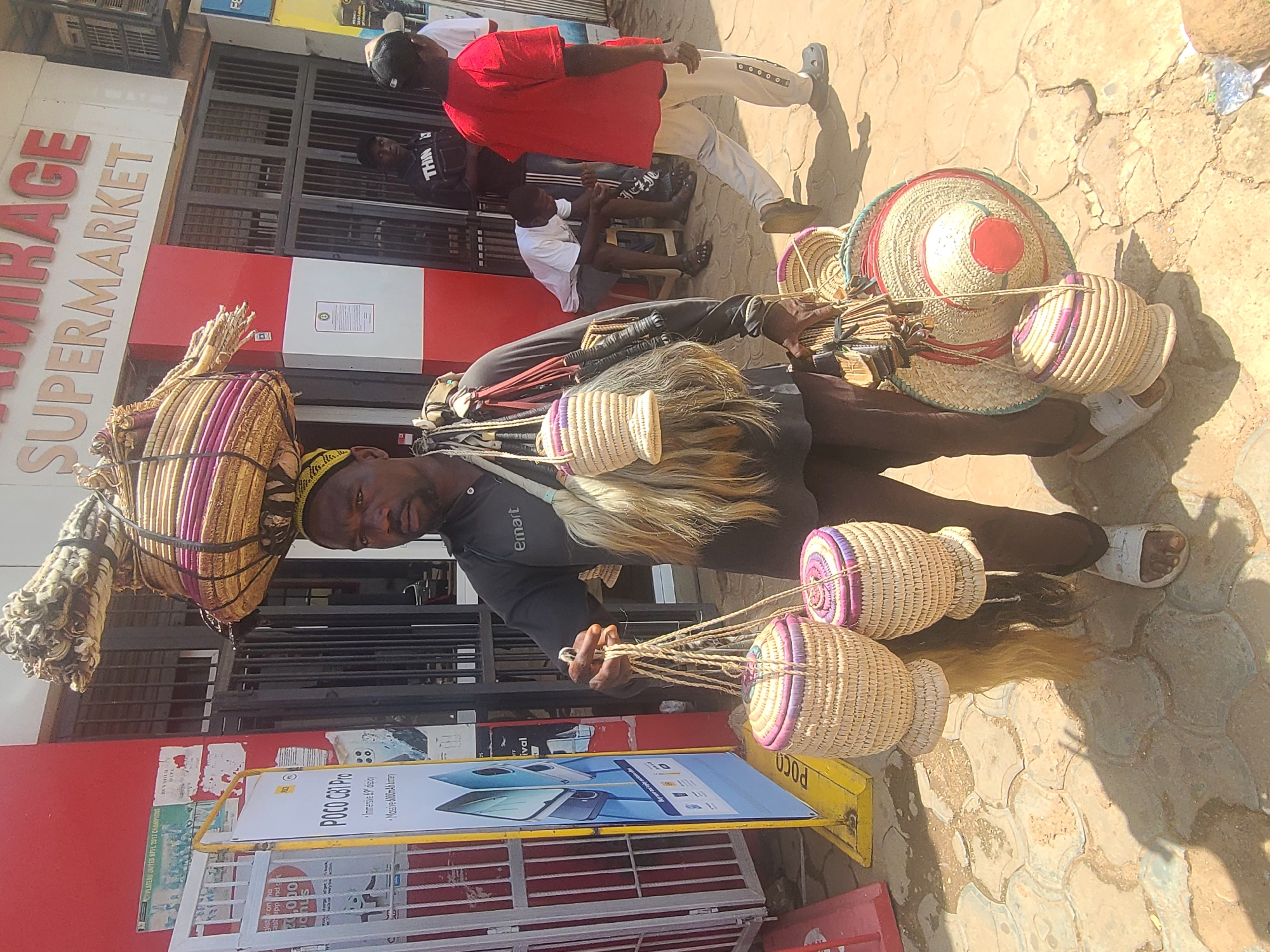A Nigerian man stands outside a supermarket in Jos, Nigeria, carrying a large woven basket on his head and several smaller woven baskets and hats in his hands. The man is wearing a dark long-sleeved shirt with "Emart" printed on the sleeve, dark pants, and white sandals. He has a dark complexion and is looking towards the camera. Behind him, the "MIRAGE SUPERMARKET" sign is visible. Other people are in the background, some near the entrance of the supermarket and others walking away. The ground is paved with interlocking stones. A yellow stand with a poster advertising a "POCO C61 Pro" phone is to the left of the man. The environment appears to be a street or market area with a building facade.