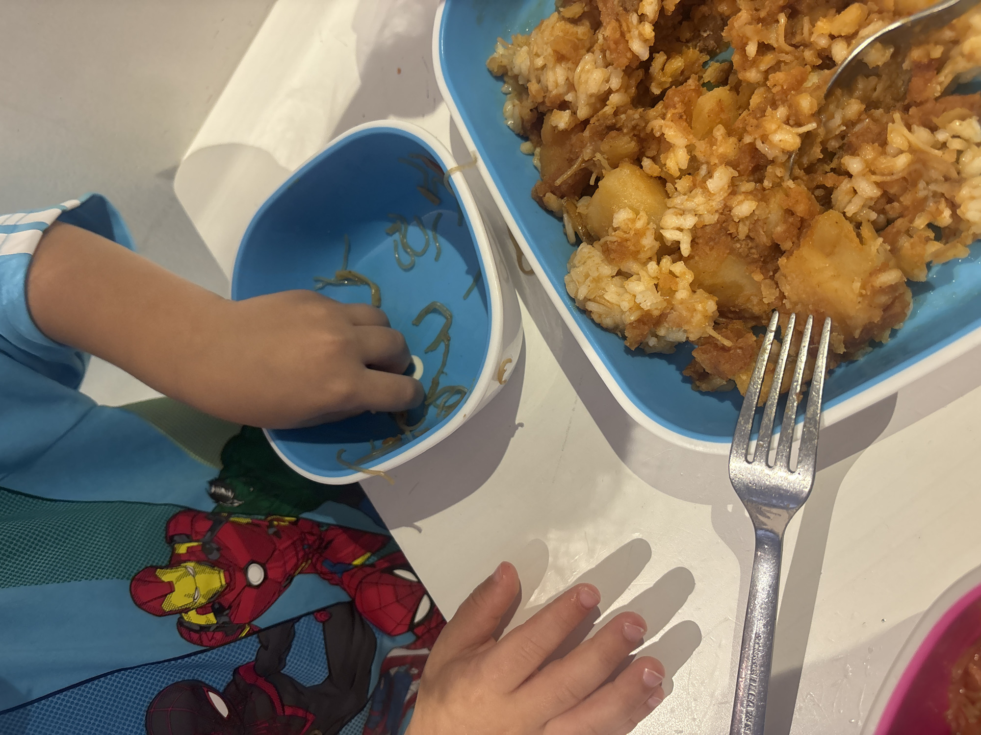 This is a close-up, eye-level shot of two children's hands reaching for food in blue plastic serving trays on a white table. The child on the left, wearing a blue t-shirt with Marvel comic characters (Spider-Man and Iron Man), has their left hand resting on the table. The child on the right has their right hand in a blue tray containing spaghetti. A second blue tray, filled with rice, potatoes, and sauce, is visible in the foreground. A silver fork is positioned over the rice dish, and a spoon is partially submerged in it. The scene appears to be indoors, possibly in a home or a casual dining setting. There is no visible indication of the specific location within Dubai, United Arab Emirates.