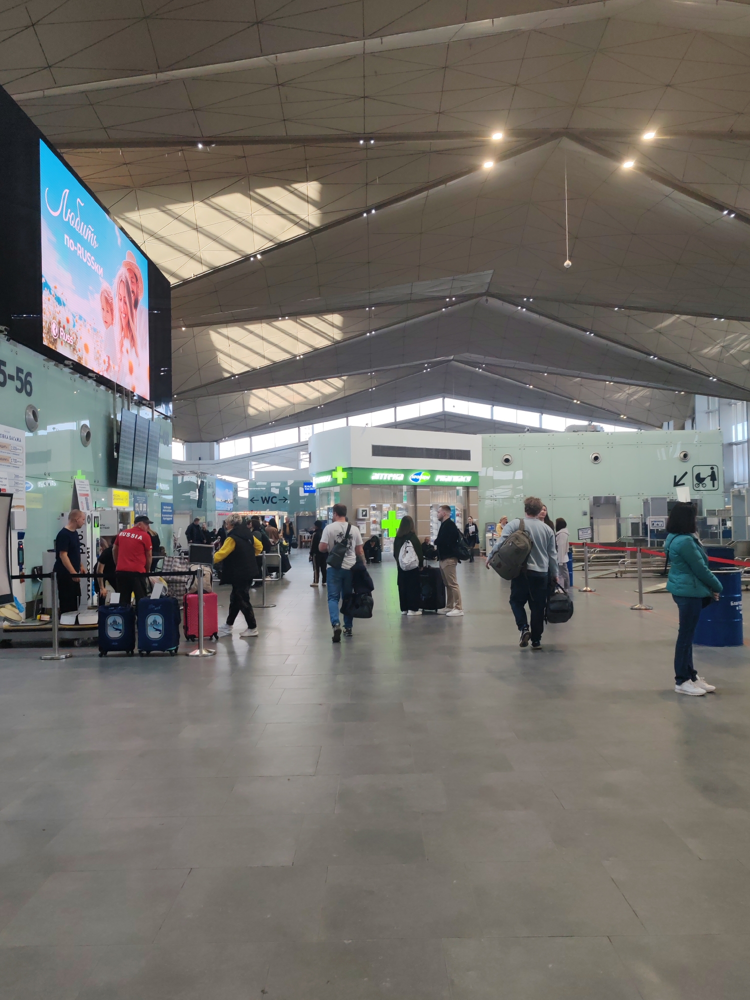 This media file captures a bright, bustling interior scene, likely within an airport terminal or a large train station in Saint Petersburg, Russia. The setting is characterized by a high, angular ceiling with triangular panels that allow natural light to filter through, casting distinct shadows. The floor is covered in large, grey tiles.
Numerous people are present, indicating a busy environment. They are seen walking, standing, and waiting, many with various forms of luggage including rolling suitcases (two blue ones featuring a distinct architectural logo, a pink one, and other black and grey cases), backpacks, and duffel bags. One man near the baggage area wears a red shirt with "RUSSIA" printed on it.
On the left, a large digital display screen shows an advertisement with the text "Любить по-RUSSKU" (Love in Russian) and a "Russ" logo, featuring an image of people in a field. Below this screen, a service counter is visible, marked "5-56" and "УПАКОВКА БАГАЖА" (Baggage Packing), with a partial price of "1700 Руб." A blue barrel or container is visible in the foreground on the right. In the background, bright green signage directs travelers to a "WC" (restroom) and an "аптека" (pharmacy), which also displays "Pharmacy" in English. Another sign with a baby carriage and person icon points towards a family facility.
The scene depicts a typical daytime travel environment, with individuals moving between different sections of the terminal.