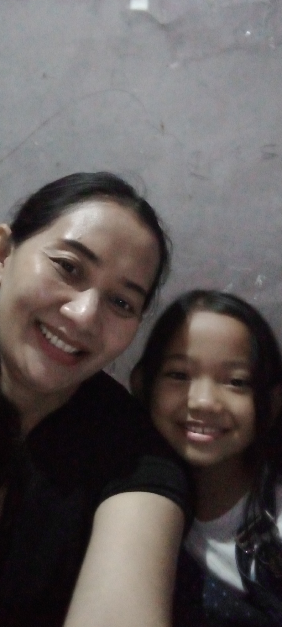 This is a close-up, self-taken photograph of two individuals. The person in the foreground on the left appears to be an adult female with dark hair pulled back and is smiling. The person on the right is a child, likely a girl, also smiling. Both are looking towards the camera. The background is a plain, textured wall, consistent with an indoor environment. There is no specific visual information that places this image within the city of Sumberwono, Indonesia; the description is limited to the visual elements present.