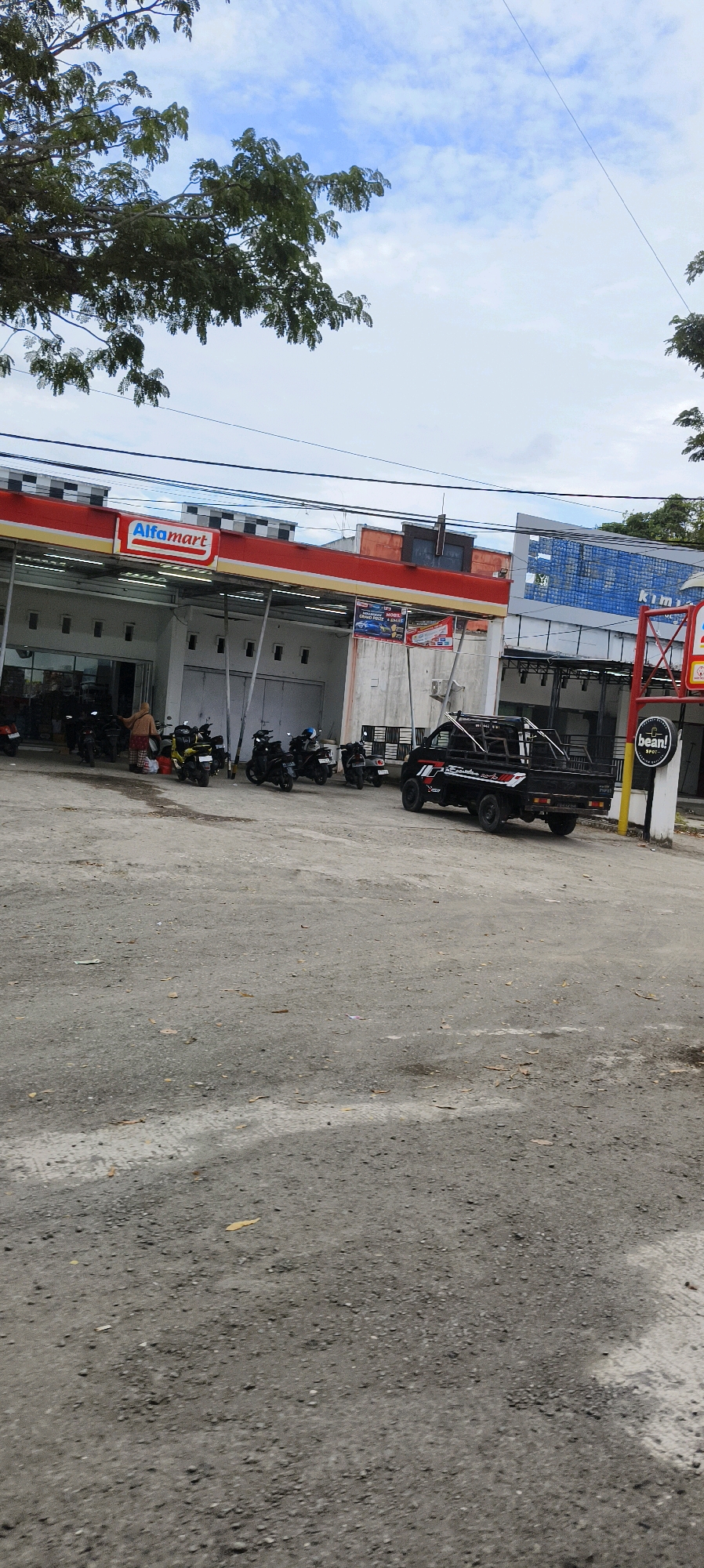 The image captures a street scene in Waisai, Indonesia, featuring an Alfamart convenience store. Several motorcycles are parked in front of the store, and a black pickup truck is positioned in the foreground. A person wearing a headscarf is standing near the motorcycles. The environment appears to be an outdoor public space with a gravelly ground surface and a cloudy sky above. The Alfamart building has a red and white awning extending over the storefront, and advertisements are displayed on the building's facade. A sign for "bean! SPOT" is visible on a yellow pole to the right of the Alfamart.