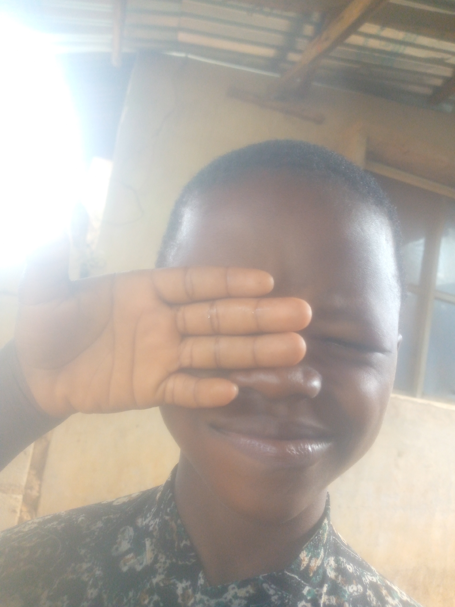 A person with dark skin is shielding their eyes from bright light with their hand. The background shows parts of a building with a window and a ceiling. The location is unknown.