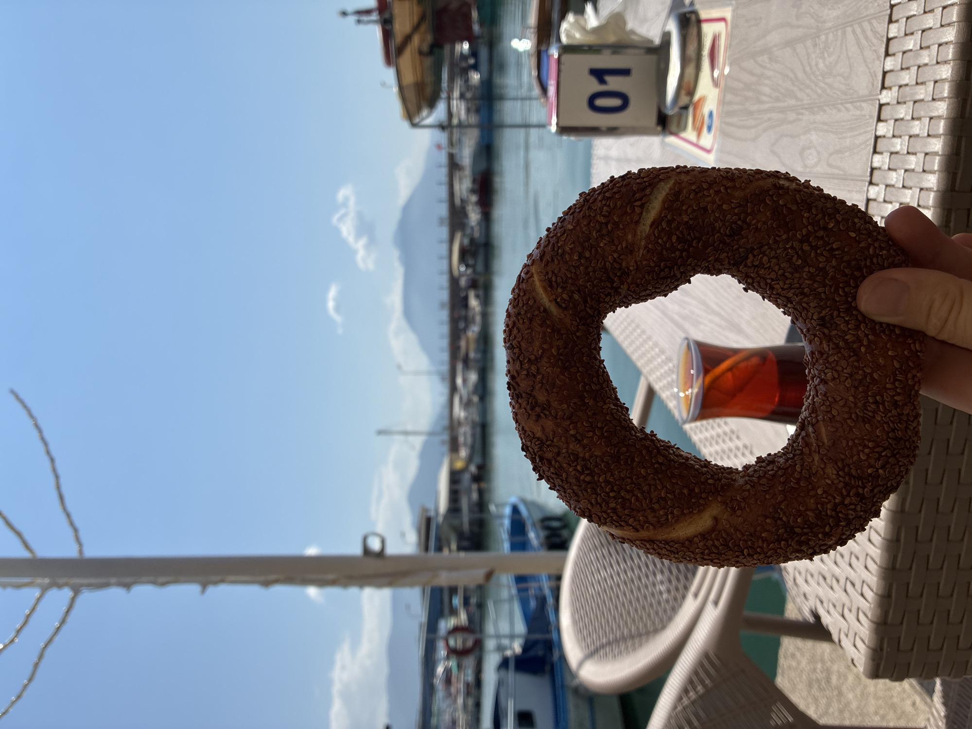 A person's hand holds a Turkish bagel, or simit, covered in sesame seeds, in the foreground. Behind the simit, a glass of dark tea sits on a woven table. In the middle ground, chairs and a table are visible, with a waterfront scene beyond. Boats are docked in the water, and buildings line the shore. In the background, mountains rise against a bright blue sky with a few scattered clouds. The overall scene suggests a relaxed meal or snack at a cafe or restaurant overlooking the harbor in Alanya, Turkey.