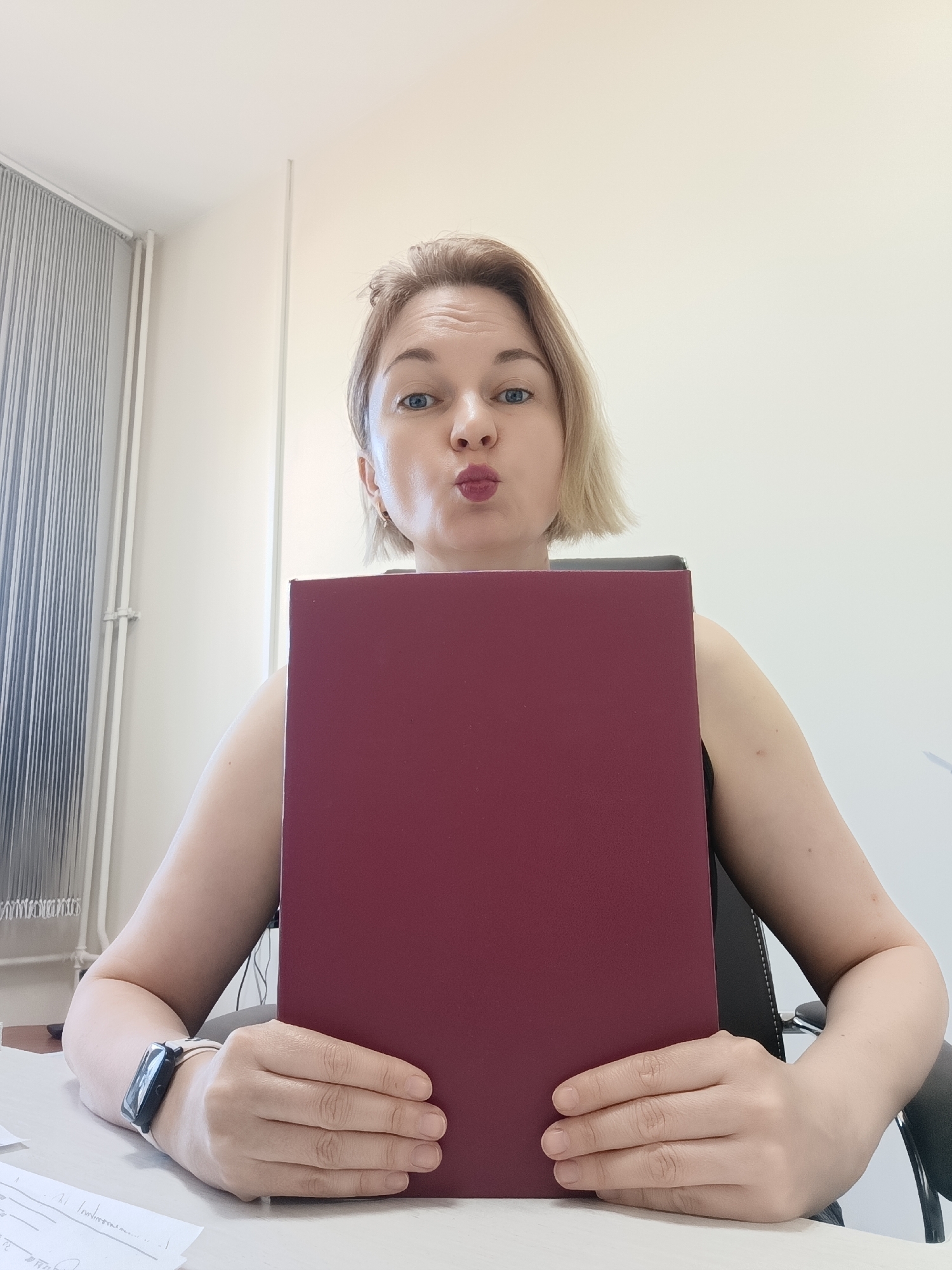 A woman with short blonde hair sits at a desk, holding a maroon document in front of her chest. She is looking directly at the camera with pursed lips. She is wearing a black smartwatch on her left wrist. The background is a white wall. There are vertical blinds to the left of the frame. The desk has papers on it. The scene is presumably indoors. The location Марковский, Russia is not detectable from the image.