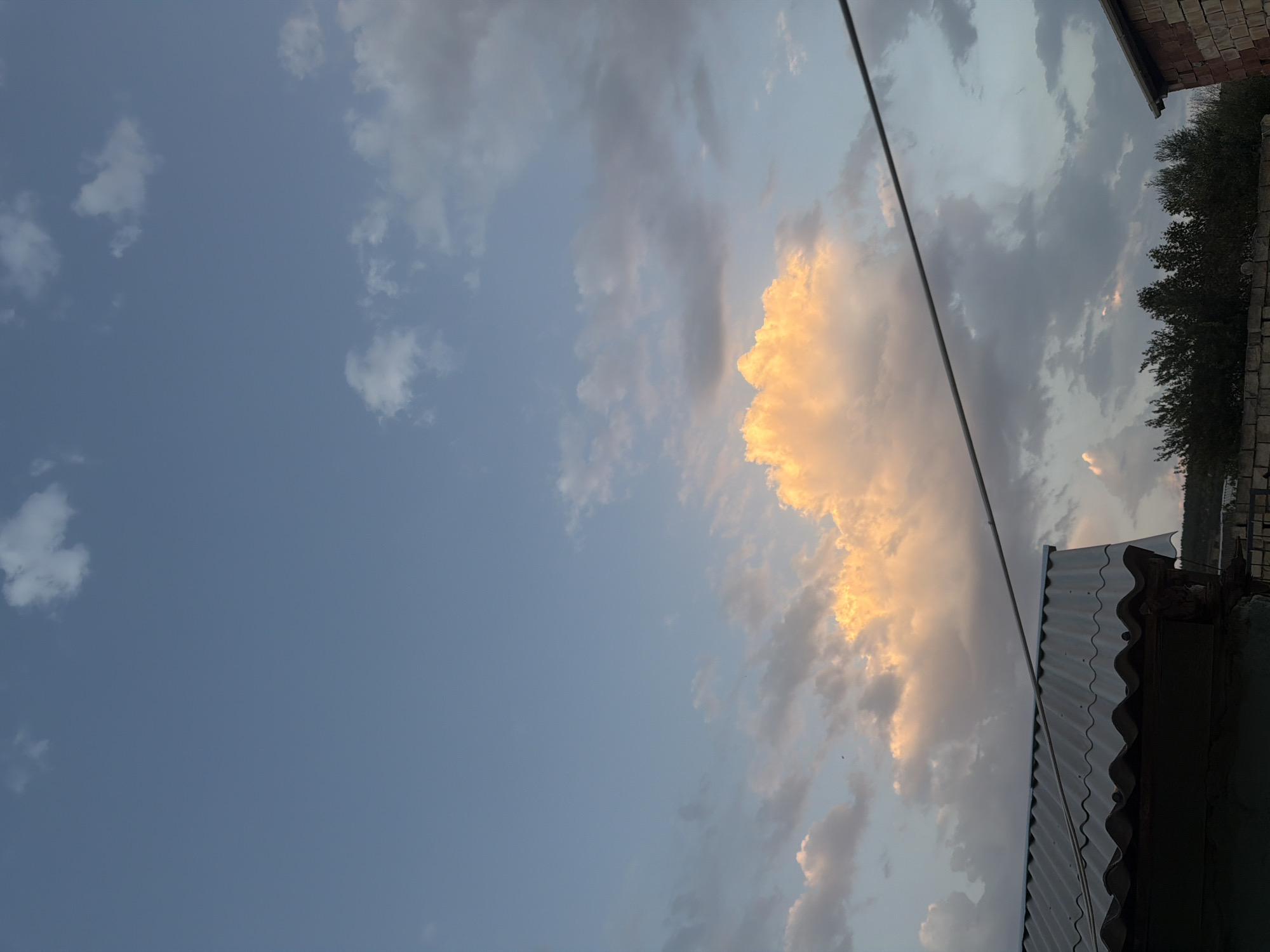 In Siazan, Azerbaijan, a twilight sky is depicted with a mixture of blue and grey clouds, some illuminated by warm, orange light. A prominent cluster of bright orange clouds is visible in the upper center of the frame. Below the sky, a corrugated metal roof of a building is seen in the lower left corner. A thin wire stretches diagonally across the image from the upper right to the lower left. In the background, a line of trees and some brick structures are visible. The scene appears to be an outdoor view from a residential area at dusk.