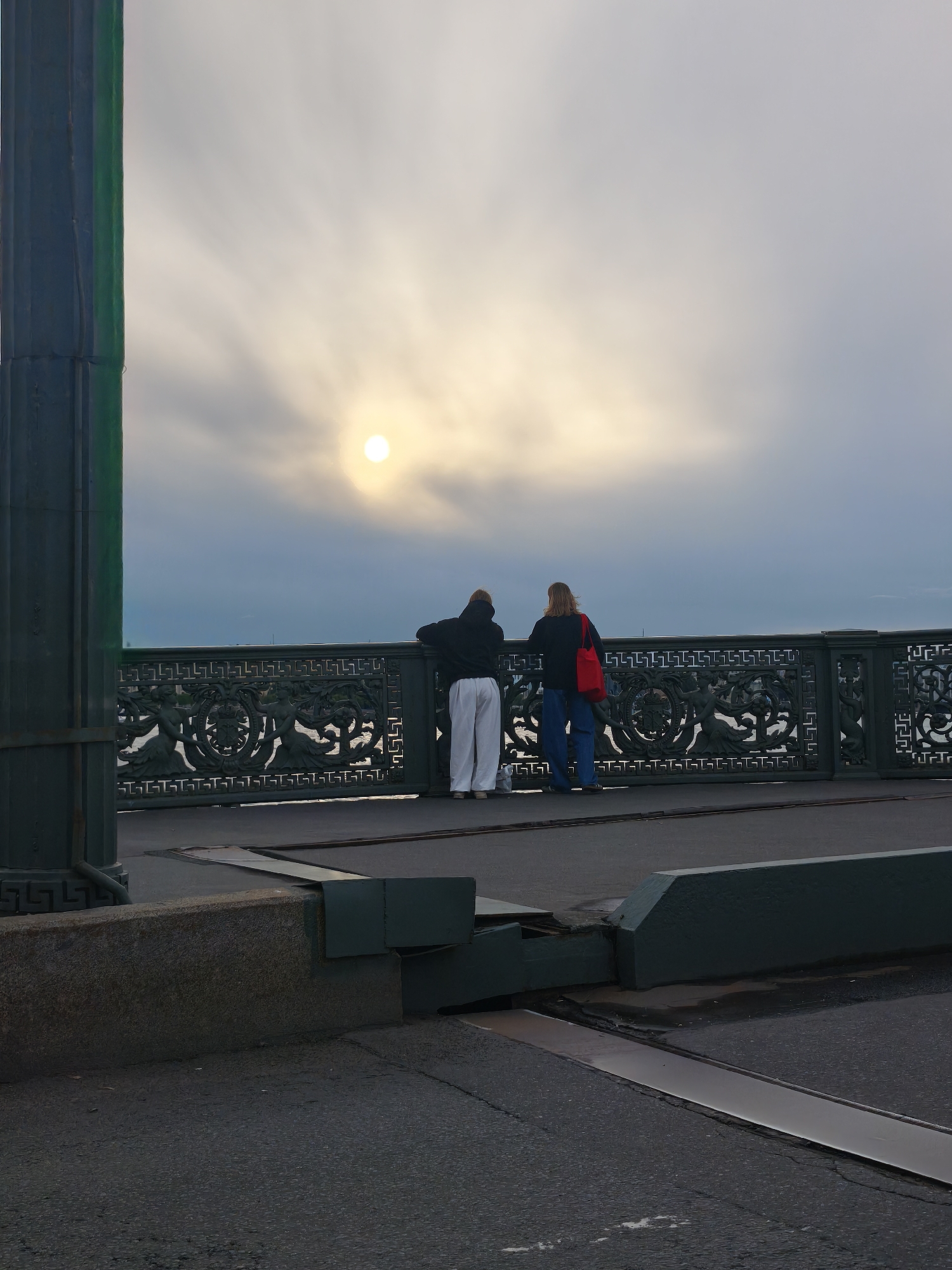 The image shows two people standing on a bridge in Saint Petersburg, Russia, looking out at the sky. The sun is visible through the clouds, suggesting it is daytime, possibly late afternoon given the soft light. The sky is overcast with wispy clouds creating a dramatic effect. The bridge railing is ornately decorated with classical motifs, hinting at the city's rich history and architecture. The foreground is dominated by the asphalt of the bridge roadway and a concrete barrier. The overall mood is one of quiet contemplation and appreciation of the scenery. The people are dressed casually, one in a dark hoodie and white pants, the other in a dark hoodie, blue jeans, and a red bag slung over their shoulder. Their postures suggest they are observing something in the distance or simply enjoying the view. The weather appears to be cool, as indicated by their clothing. No visible text is present in the image.
