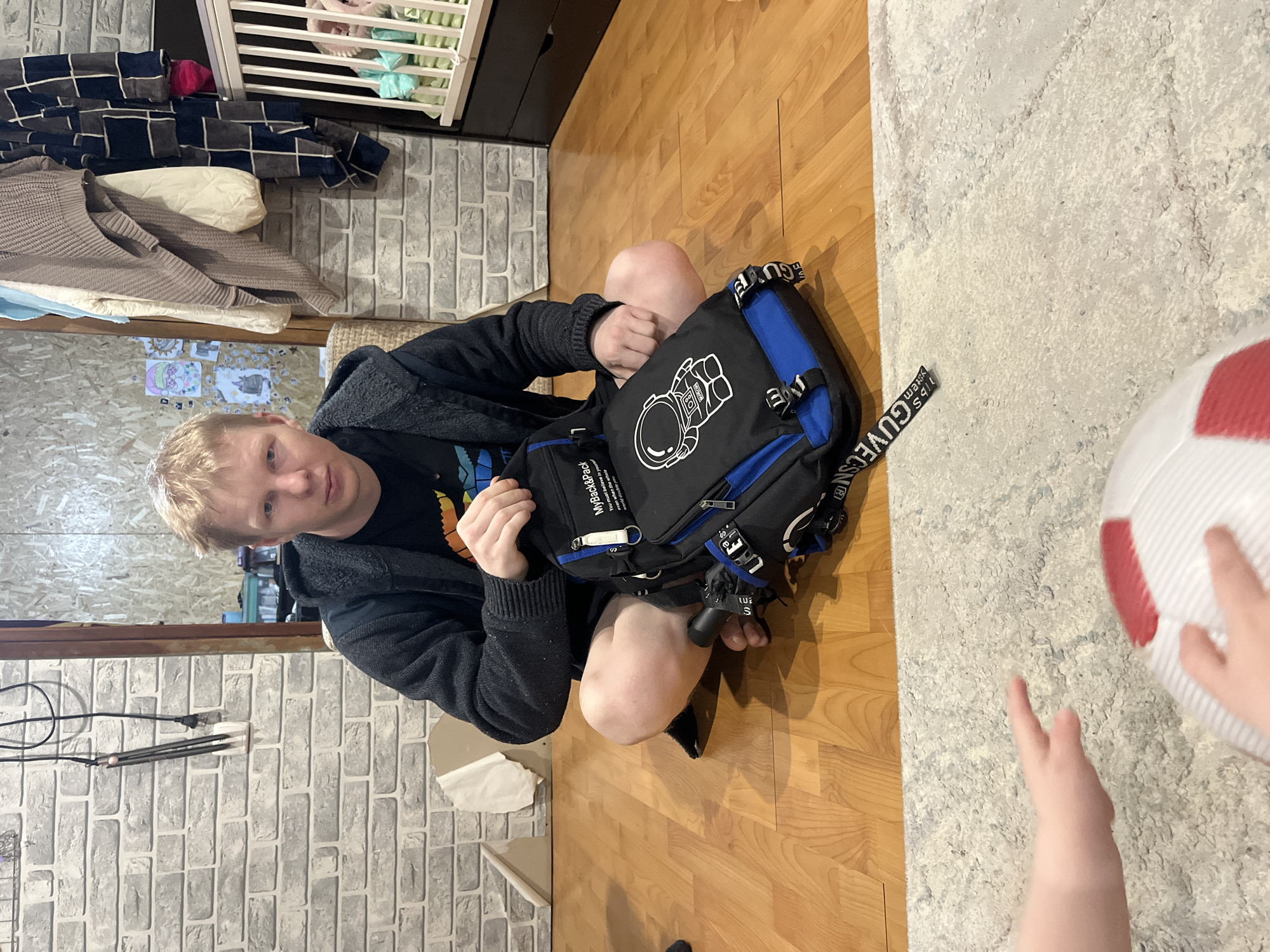 A high-angle shot reveals a person sitting cross-legged on a wooden floor, examining a black and blue backpack. The backpack features a white astronaut design. The person is wearing a dark hoodie over a t-shirt. To the person's right, a child's hand and a red and white ball are visible on a light-colored rug. The background includes a faux brick wall, a baby crib, and other household items. The location is Vesyoloye, Russia.