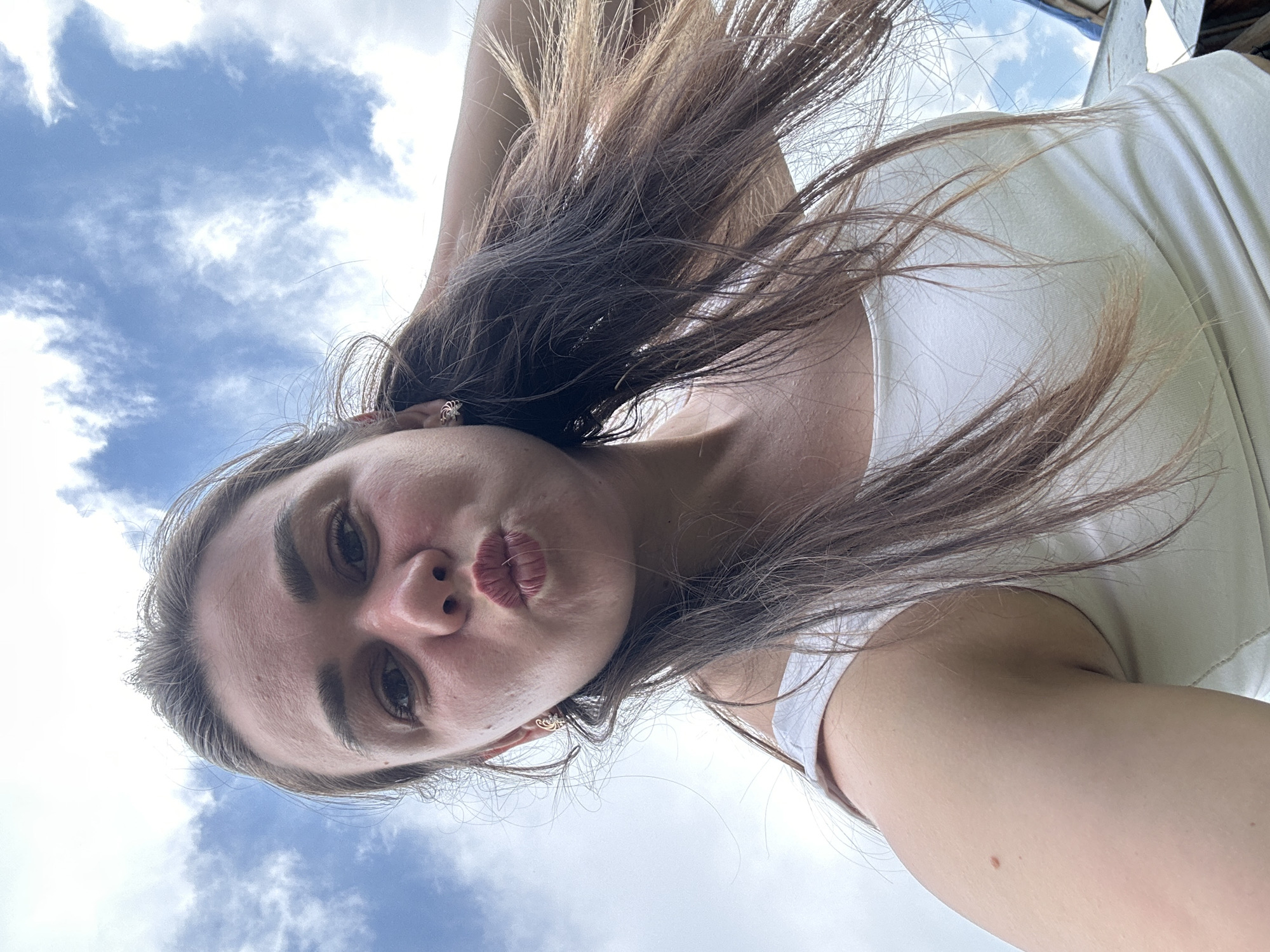A young woman with brown hair is taking a selfie outdoors. She is wearing a white tank top and has her hair over one shoulder. The sky with white clouds is visible in the background. The image appears to be taken from a low angle, looking up at the woman. The location is likely outdoors, in a residential area of Chaikovsky, Russia, given the context.