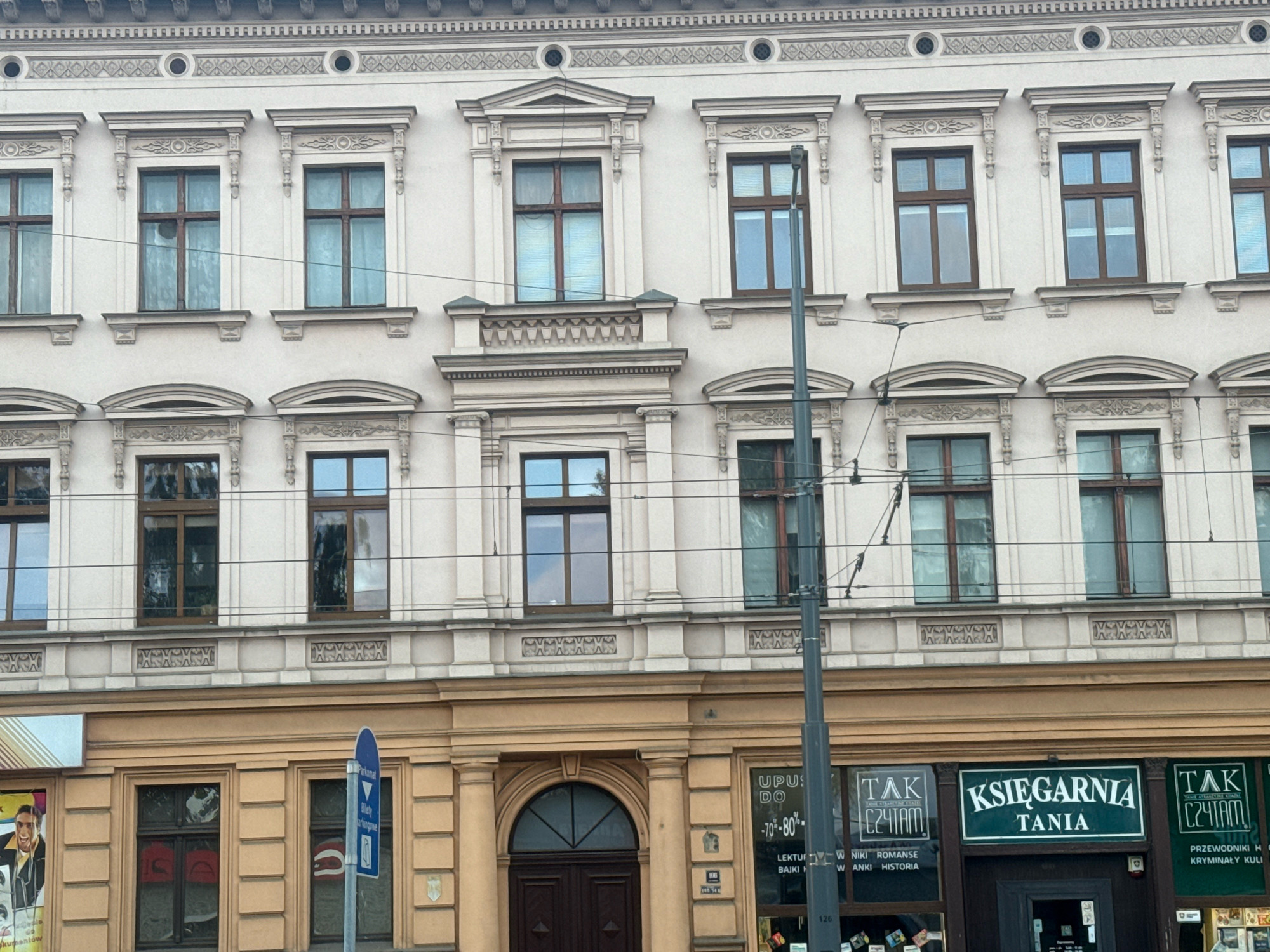 This is a medium-shot, eye-level exterior photograph of a building's facade in Gorzow, Poland. The building has a light-colored, ornate facade with multiple rows of windows and decorative elements. The upper floors feature numerous rectangular windows with dark frames, each adorned with intricate architectural detailing above and below. Some of these windows have blinds drawn, while others show reflections of the sky and surrounding environment. A thin, grey pole, likely a streetlamp or utility pole, bisects the frame vertically on the right side.
The lower floor of the building is a different color, a warm, light brown, and features storefronts. The central storefront displays a green sign with white lettering that reads "KSIĘGARNIA TANIA" (Bookstore Cheap). To the left and right of this central sign are other storefronts with signs that appear to be for a bookstore as well, with text including "TAK CZYTAM" (Yes, I read). One of these storefronts has a large poster featuring a smiling man in a suit. A blue directional sign is visible on a pole near the center of the image, indicating parking. The lower part of the building exhibits a more minimalist design compared to the upper floors, with simple columns flanking the entrance and window frames. Electrical wires are visible strung across the facade. The overall impression is an urban street scene in front of a historic building with commercial establishments on the ground floor.