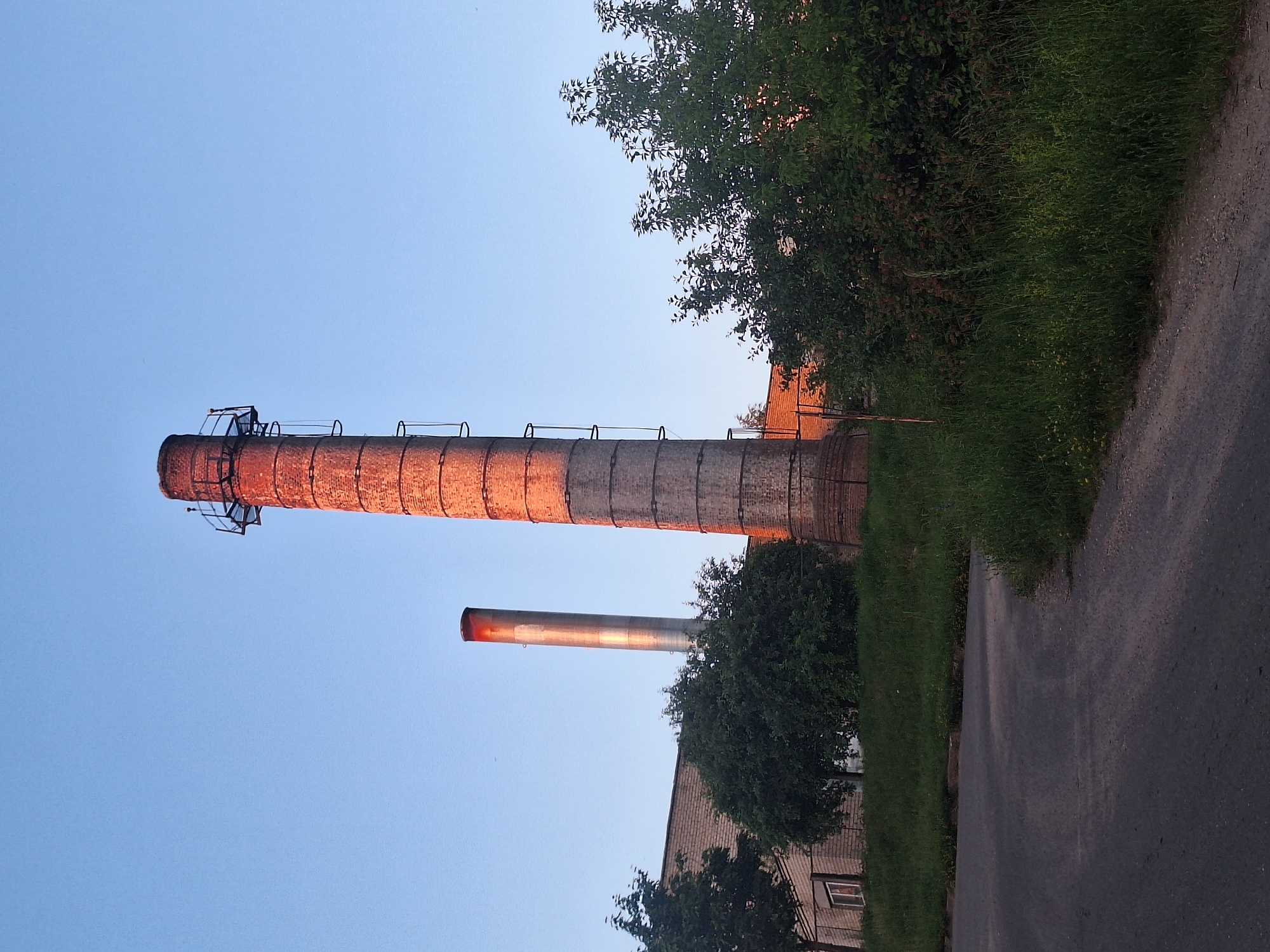 The image depicts an outdoor scene with two industrial chimneys against a clear blue sky, illuminated by the warm glow of sunset. The larger chimney, made of brick, stands on the right, with a metal ladder affixed to its side. To its left, a taller, slimmer metal chimney emits a faint red glow from its top. In the mid-ground, a low-rise brick building with a window is visible, partially obscured by dense green foliage and trees. A paved road curves through the foreground, bordered by grassy slopes. The scene appears to be located on the outskirts of the city of Капыльскі сельскі Савет, Belarus. There are no people visible in the image.
