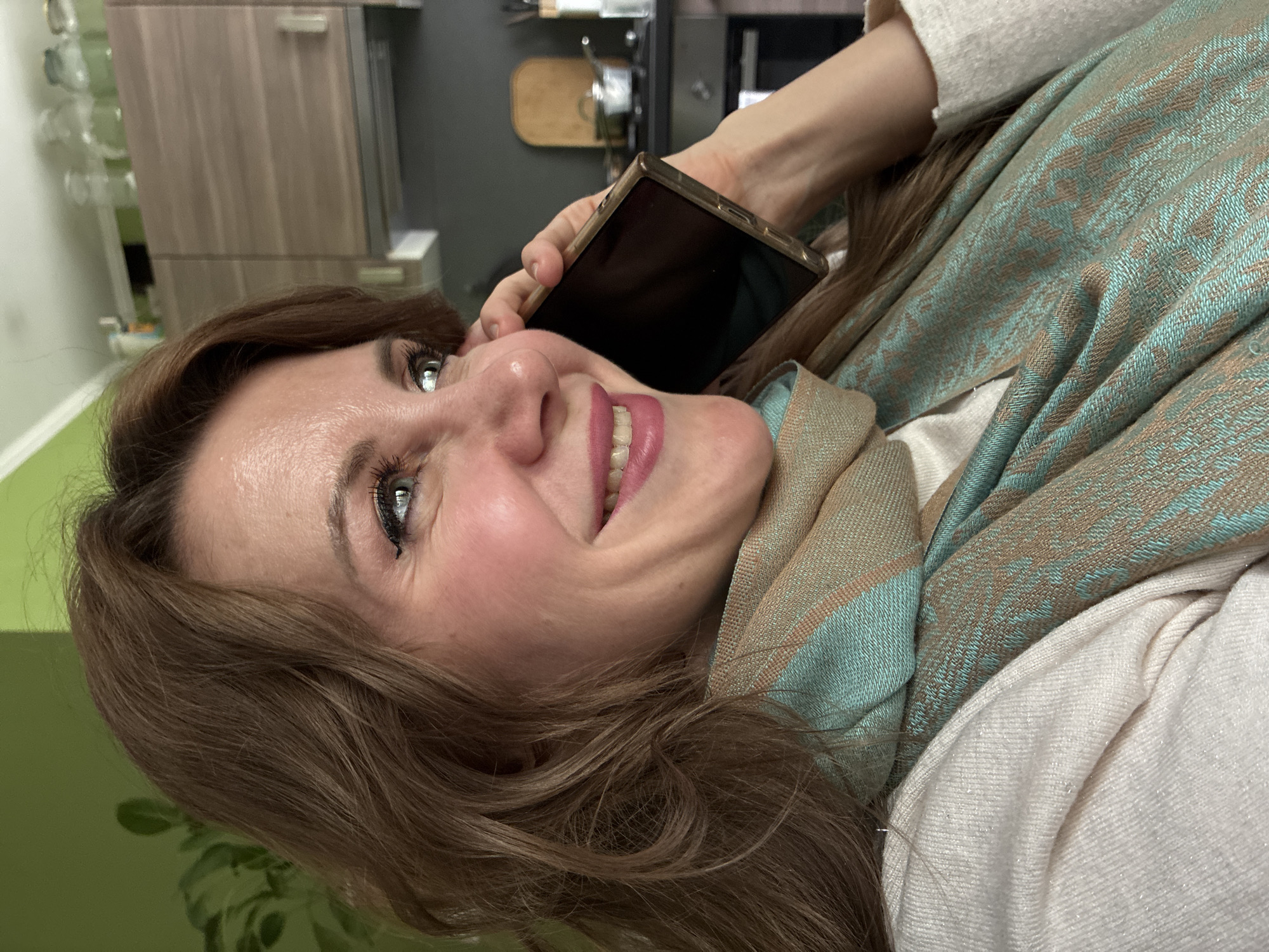 A woman with light brown hair is smiling and holding a black smartphone to her ear. She is wearing a white top and a turquoise and beige scarf. The background includes a kitchen with wooden cabinets and appliances. It is not possible to confirm this photo was taken in Moscow, Russia.