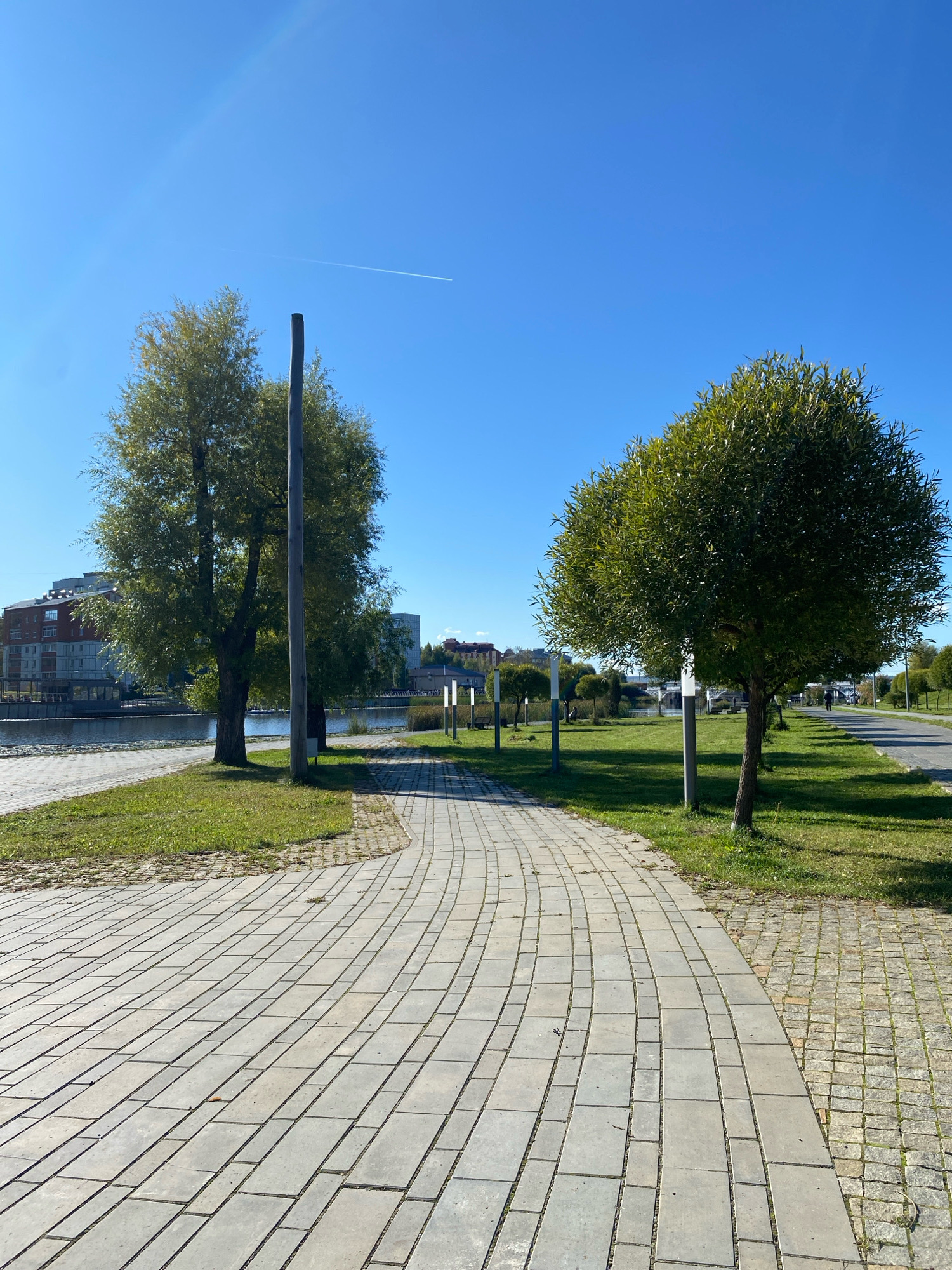 The image depicts a paved walkway curving through a park-like setting on a clear, sunny day. The walkway is constructed from light grey rectangular pavers laid in a staggered pattern, with a narrower section of smaller, square pavers to the right. Lush green grass borders the walkway on both sides.
On the left side of the walkway, near the foreground, are two large trees with full, leafy canopies. A tall, rough-hewn wooden pole stands between them. Beyond the trees, a body of water, likely a river or canal, is visible, with buildings lining its far bank. These buildings appear to be modern, multi-story structures with reddish-brown and grey facades.
On the right side of the walkway, several modern lampposts with white cylindrical lights are spaced along the path. Further in the distance, a single person is walking along a paved road that runs parallel to the walkway. The sky is a vibrant, clear blue with a faint white contrail from an airplane visible high above.
The scene suggests a public recreational area within an urban environment. While the prompt specifies Votkinsk, Russia, there are no overt visual cues in the image itself that definitively confirm this location. The architecture of the buildings and the general style of the park are not exclusively tied to any single Russian city without further context.