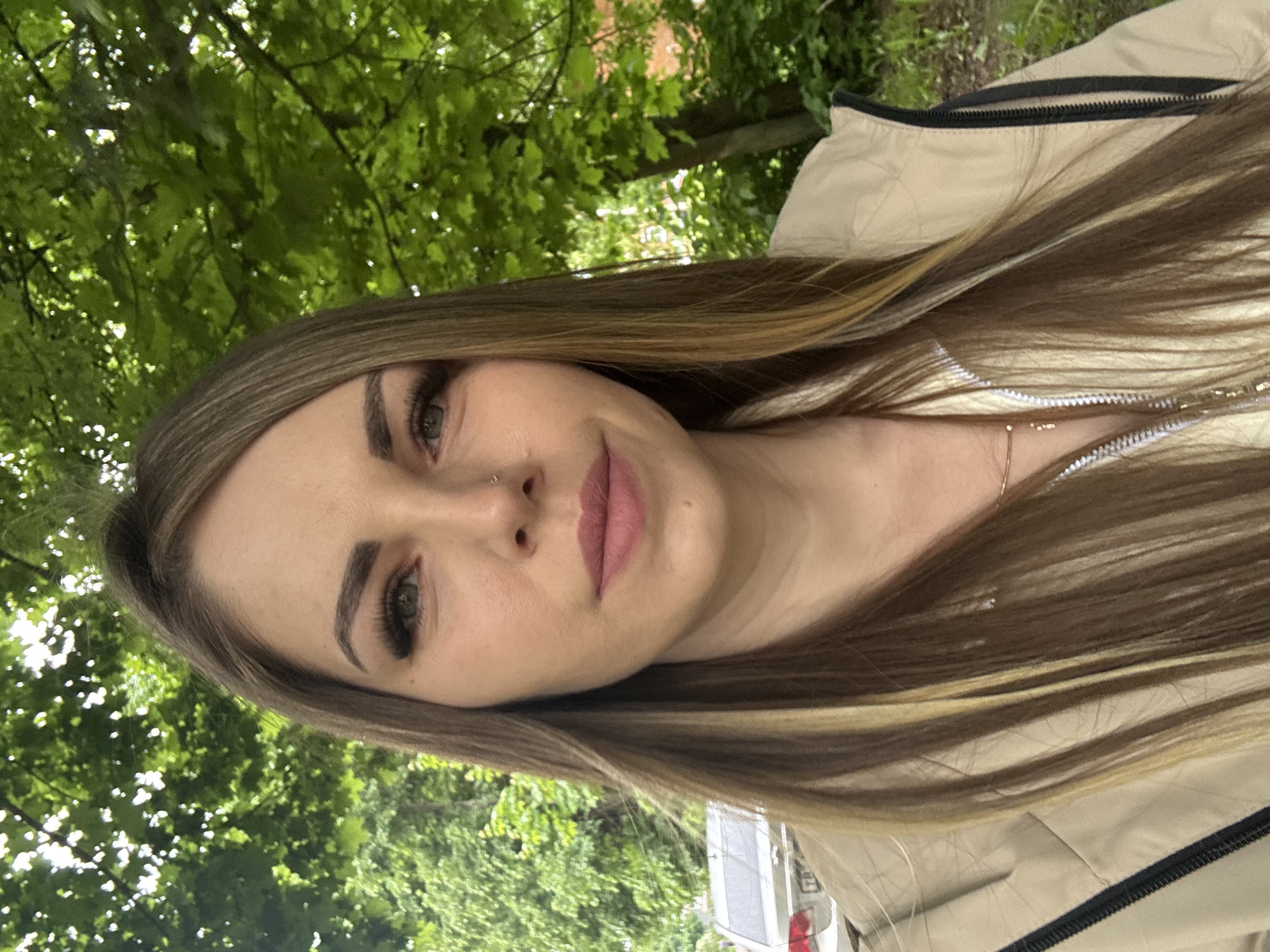 A woman is depicted outdoors, likely in a park or wooded area due to the abundant green foliage of trees in the background. The subject is wearing a light-colored jacket and a thin necklace. She has long, highlighted hair and appears to be wearing makeup. The image does not contain sufficient information to place it in the city of Shevlyakovo, Russia, or determine if the image was even taken there. There are no actions, interactions, or other people visible.