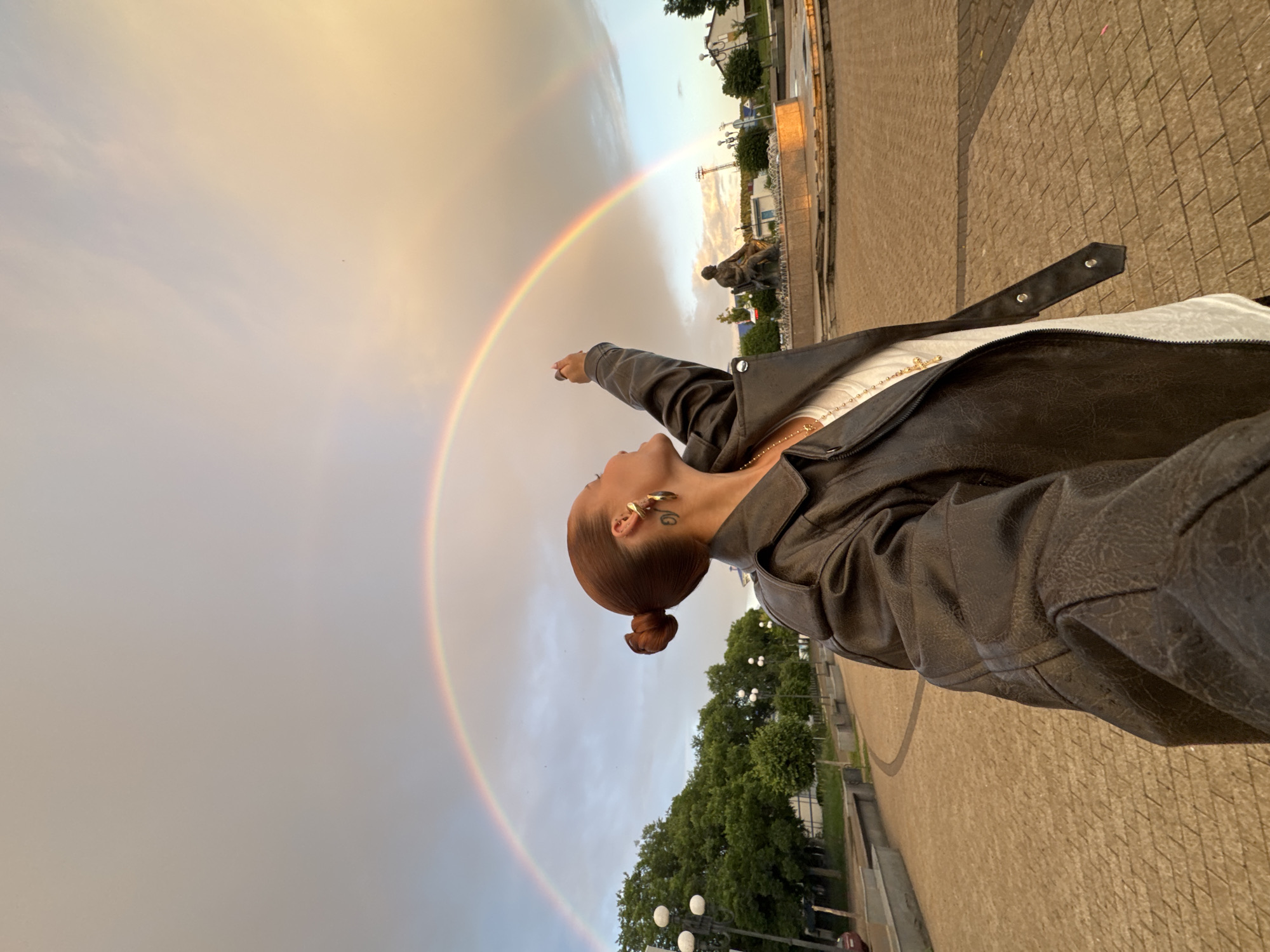 The media file captures a person posing in front of a double rainbow under a bright sky. The person, whose back is to the camera, is wearing a black leather jacket over a white top. They are looking up and to their left, with their left arm extended upwards, pointing towards the rainbow. The scene is set outdoors on a paved area, likely a public square or park, with trees and buildings visible in the background. A statue is also present in the mid-ground. The lighting suggests it is either early morning or late afternoon, with the warm tones of the sky indicating the presence of sunlight filtering through clouds, which also facilitated the formation of the rainbow. The weather appears to be fair, with the rainbow signifying recent or ongoing rain. The overall mood is positive and aspirational, evoked by the vibrant rainbow and the person's gesture. No text is visible in the media file. The location context provided is Chaikovsky, Russia.