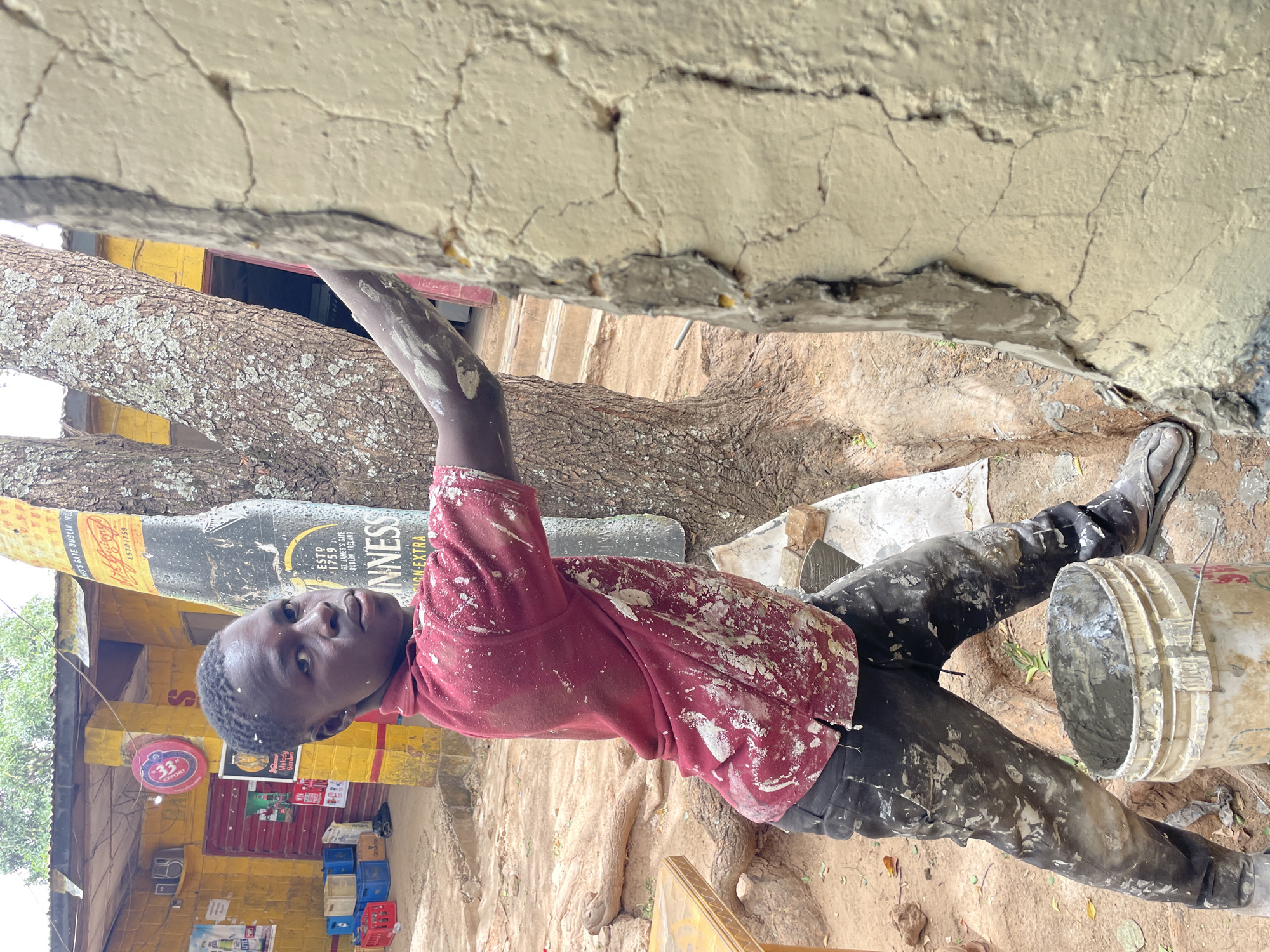 A man plasters a wall in Jos, Nigeria. The man, covered in plaster, wears a red shirt and black pants. He kneels on the ground beside a bucket of plaster. Behind him is a yellow building with a Guinness advertisement.