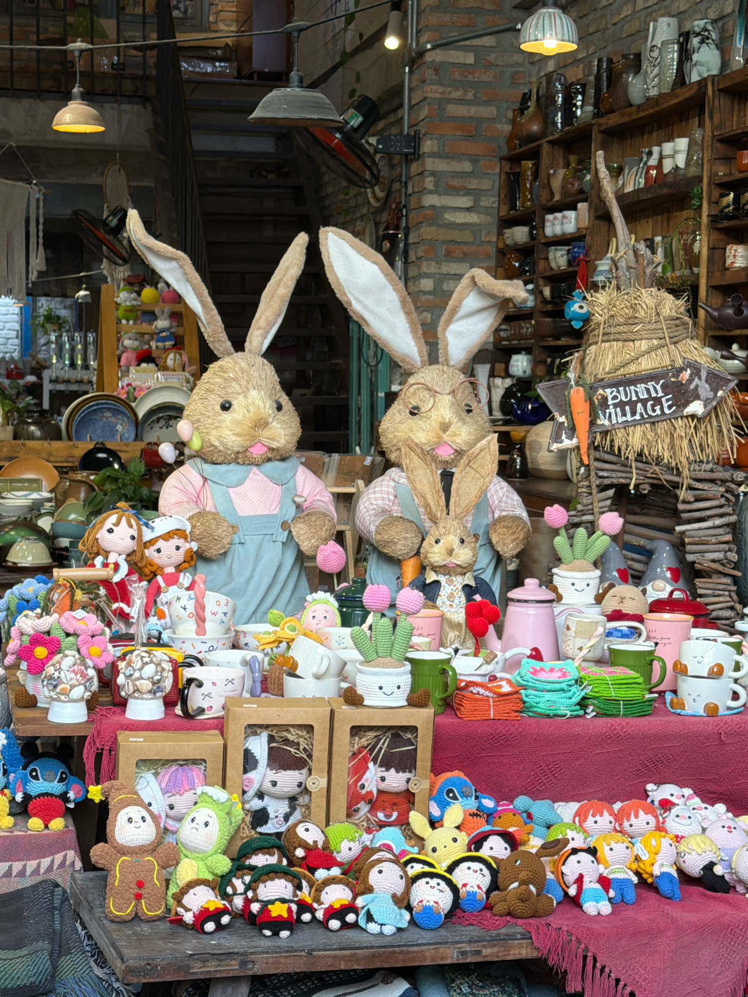 This image depicts an outdoor stall in Nha Trang, Vietnam, likely a market or street vendor setting, filled with a variety of handcrafted items, primarily for Easter or spring themes. Two large plush bunnies, dressed in overalls and pastel shirts, are the centerpiece. In front of them, a collection of crocheted dolls, small plush toys, and ceramic mugs are displayed on a red fabric-covered surface. Smaller crocheted bunnies and other figures are scattered throughout. The background reveals shelves stocked with pottery and decorative items, suggesting a craft store or a stall specializing in handmade goods. The overall impression is one of a colorful and whimsical display aimed at attracting customers.