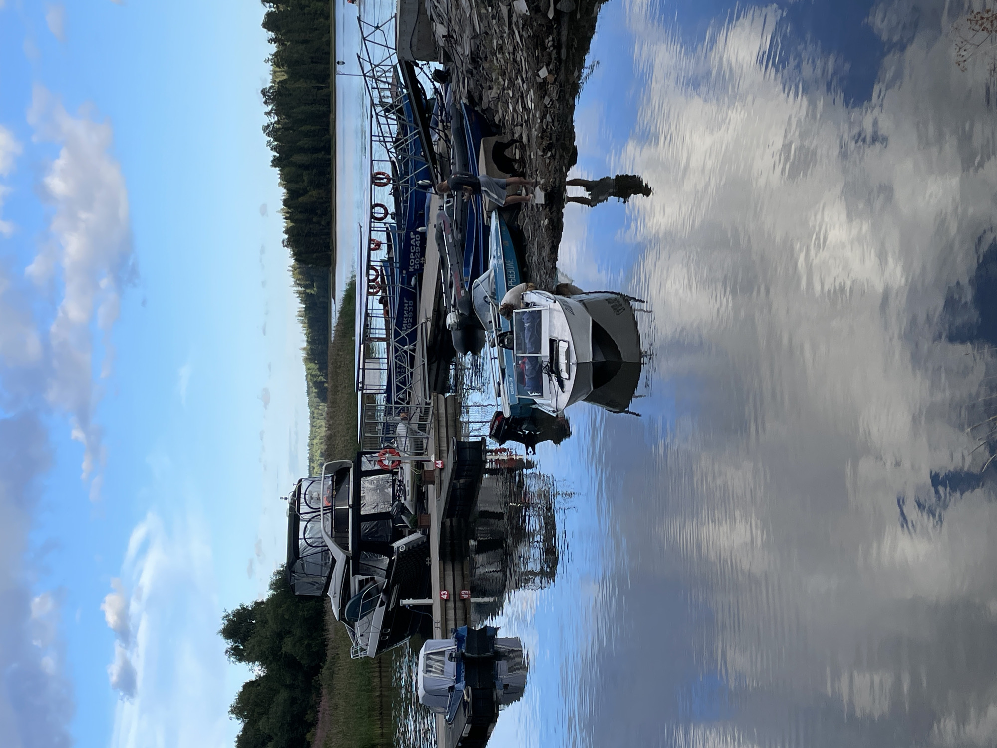 In the city of Berozovo, Russia, a serene waterscape unfolds, featuring a collection of boats docked along a pier. The sky, adorned with scattered clouds, casts reflections on the calm water, creating a mirror-like effect. Among the docked vessels, a prominent large boat with multiple levels and a covered top occupies a central position. Smaller boats, some with protective covers, are also visible. On the pier, a man stands, holding a phone, while a dog is nearby. The environment is characterized by the presence of trees in the background, indicating a natural setting. The scene is captured from a slightly elevated perspective, offering a comprehensive view of the boats and their surroundings.