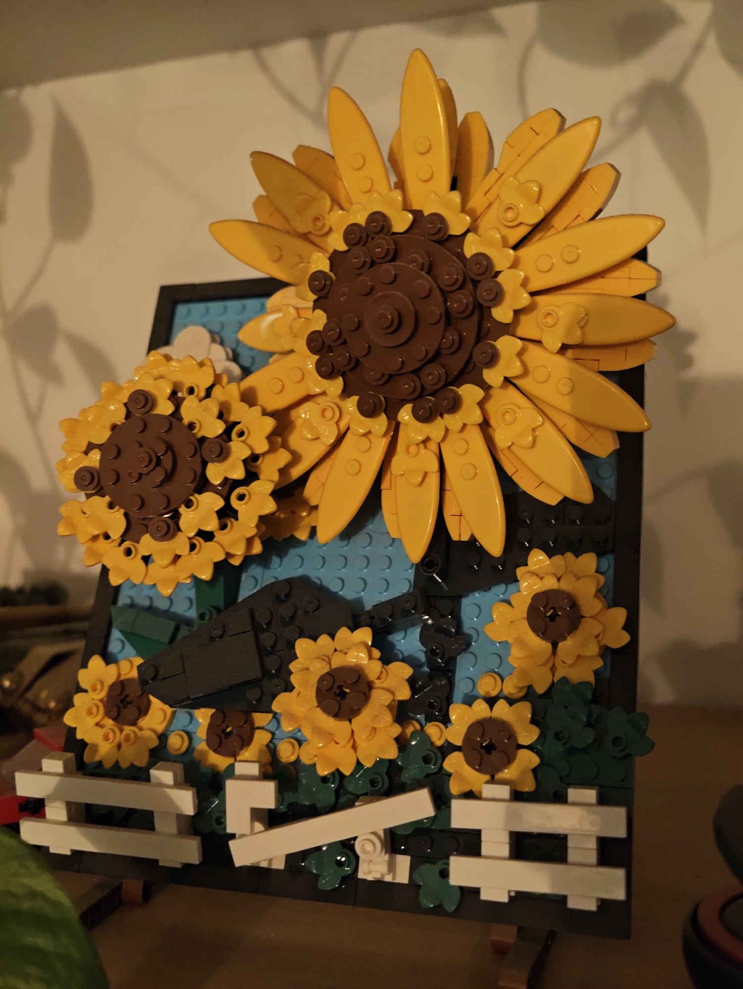 The image depicts a LEGO model of sunflowers displayed on a dark frame against a light-colored wall, which has subtle shadows cast upon it, suggesting indoor lighting. The model features several sunflowers of varying sizes, constructed with yellow and brown LEGO bricks. The larger sunflower dominates the upper portion of the frame, with its petals radiating outwards. Smaller sunflowers are arranged below and to the left. A white LEGO fence runs along the bottom of the model. The background within the frame consists of blue LEGO bricks, possibly representing a sky or body of water. The overall scene is a static display of a creative LEGO build. There is no indication of the city of Petah Tikva, Israel, in the image or its contents.