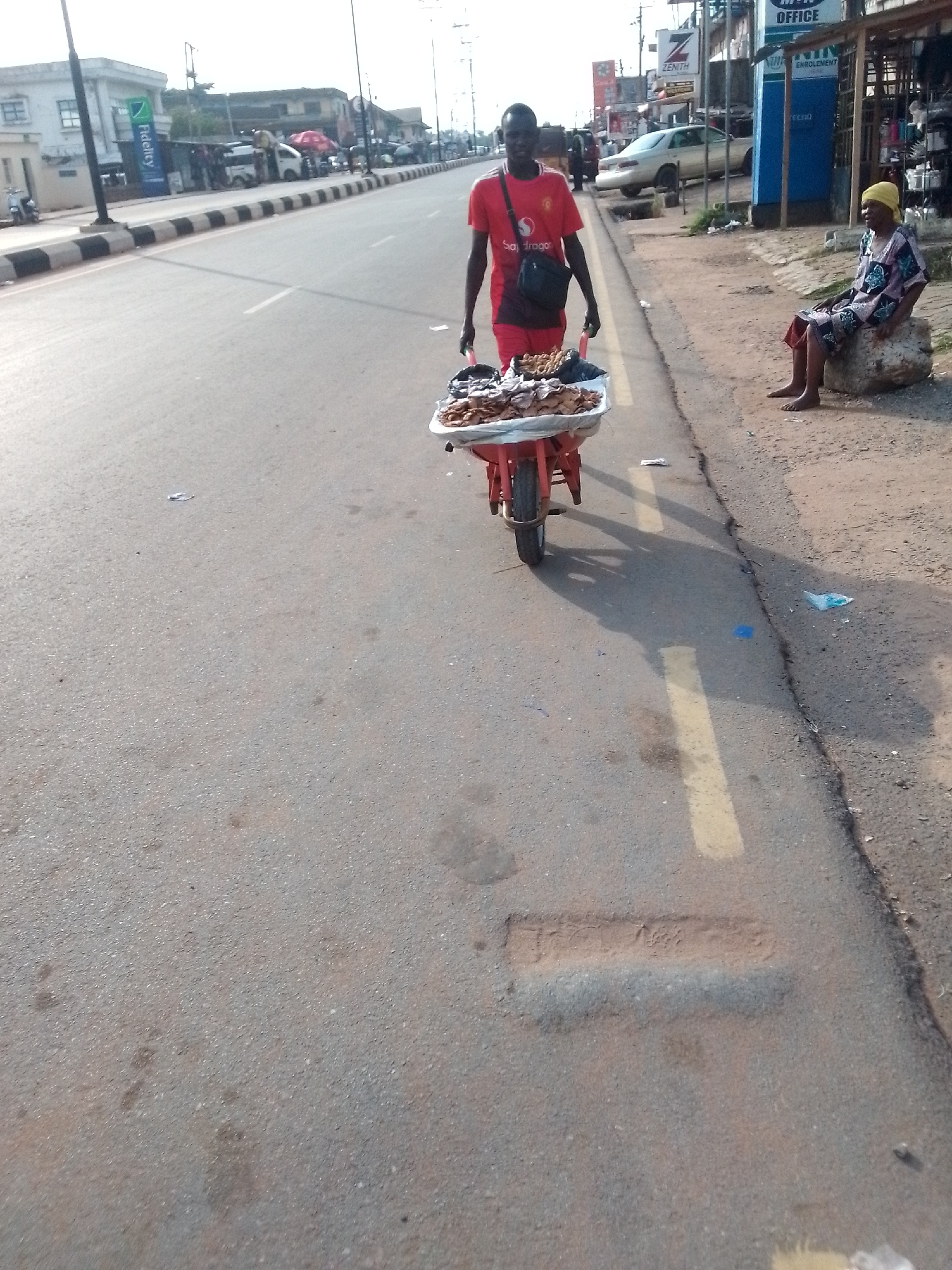 The image depicts a street scene in Orlu, Nigeria. A man in a red t-shirt and shorts is pulling a red wheelbarrow filled with food items. He is walking on the paved road, near the edge. An older woman wearing a yellow headscarf and patterned clothing is sitting on a rock on the side of the road. In the background, there are buildings, shops, and parked vehicles. A bank (Fidelity) logo is visible in the background. The road has a faded yellow line marking. The overall light suggests it is daytime.