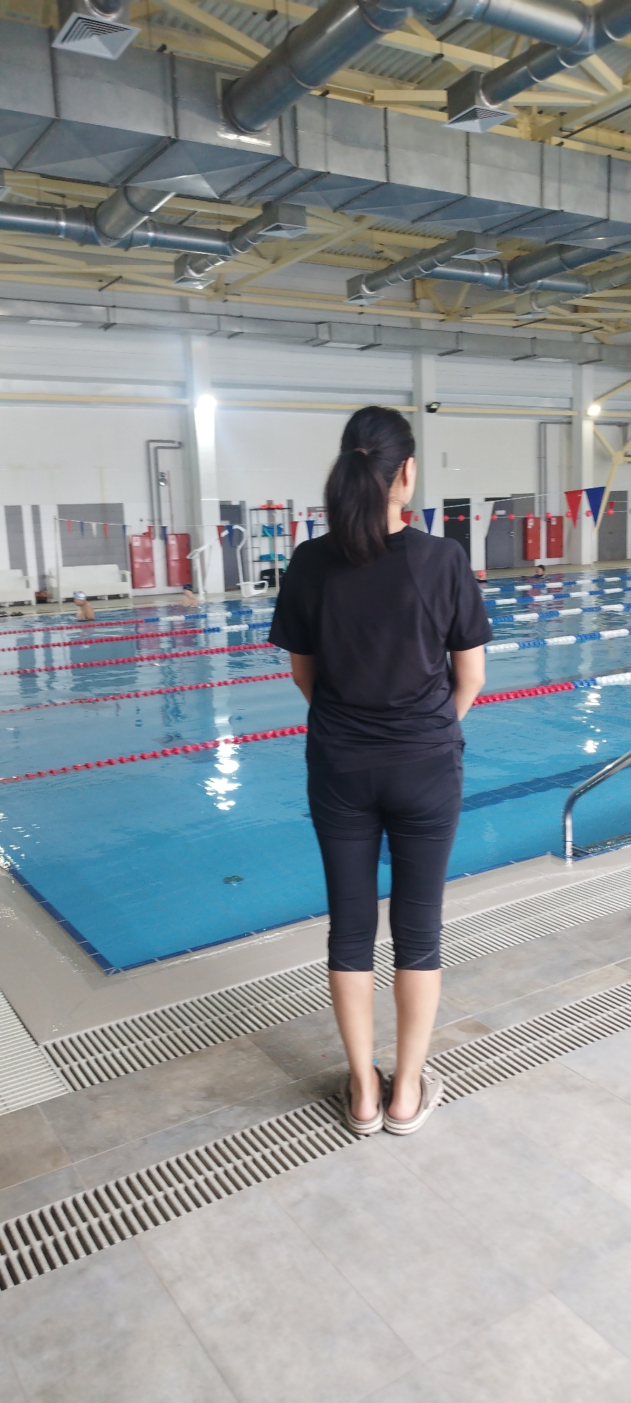 A woman stands with her back to the camera, observing a swimming pool. She wears a black T-shirt, black capri pants, and open-backed shoes. The pool has lane markers and is surrounded by tiled flooring with drainage grates. The background shows a large indoor space with a white wall and rows of competition flags. Metal ductwork and vents are visible on the ceiling. The location is in Yakutsk, Russia.