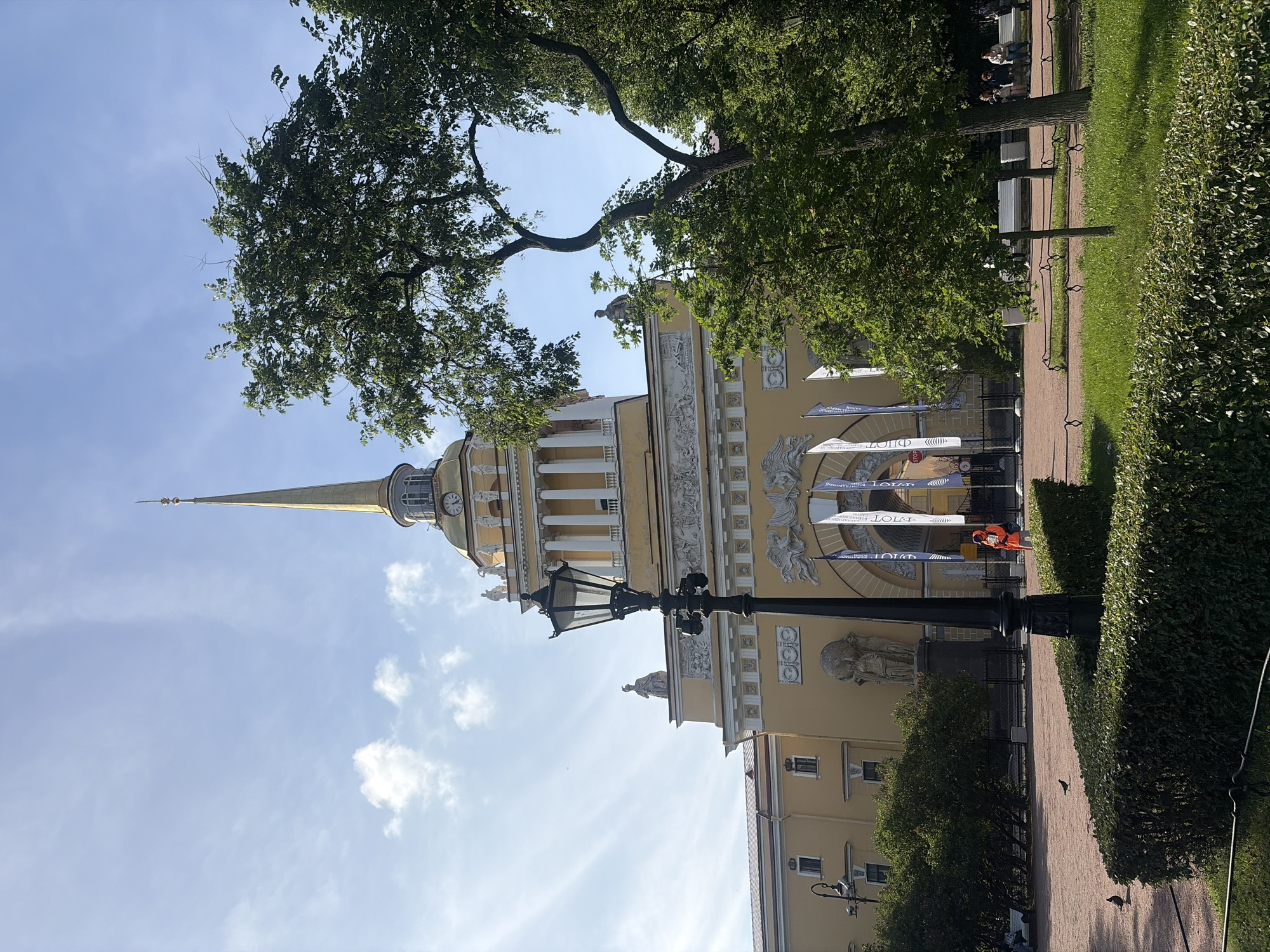 The image shows the Admiralty Building in Saint Petersburg, Russia. The building is a prominent yellow structure with a tall, golden spire topped with a weathervane. The spire is visible against a bright blue sky with scattered white clouds. In the foreground, there is a black lamppost and well-maintained green hedges and grass.
Several people are visible in the scene. A person in an orange jacket is walking near the entrance of the building. In the middle ground, some individuals are sitting on benches in a park-like area.
The Admiralty Building itself features classical architectural elements, including columns, statues, and decorative reliefs. Several white flags with blue lettering are displayed in front of the building's main entrance. Lush green trees with dense foliage frame the right side of the image, partially obscuring the building. The ground in the foreground is covered with a light pinkish gravel.