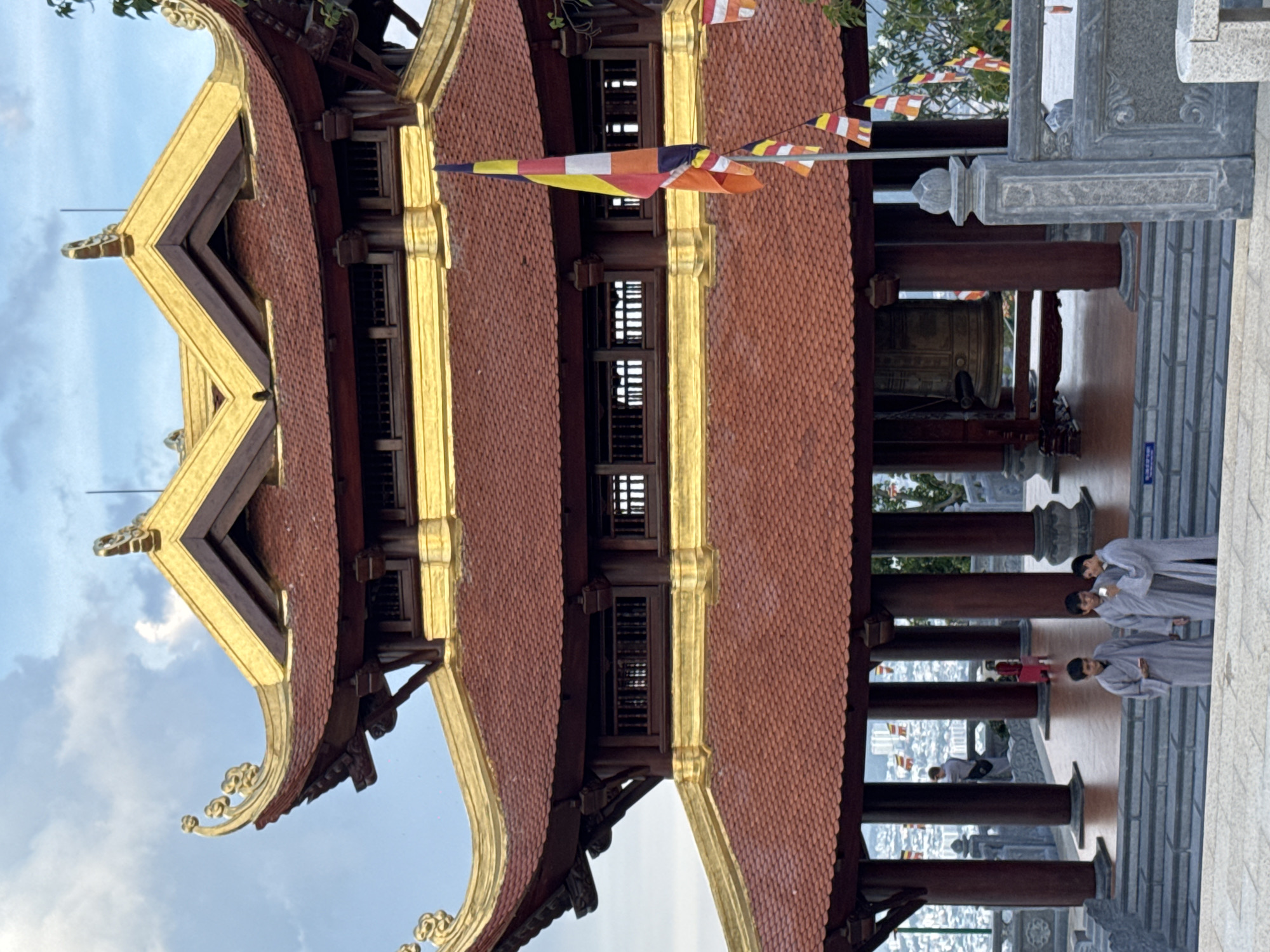 A multi-tiered pagoda, adorned with red tiled roofs and gold trim, stands tall against a cloudy sky. The pagoda features intricate wooden carvings and supports large, dark red pillars. At the base of the pagoda, a large bronze bell is visible, suspended within a sheltered area. Several Buddhist flags in various colors, including yellow, orange, white, and blue, flutter from a flagpole in front of the pagoda.
In the foreground, three young men, dressed in light grey monastic robes, stand on a wide stone staircase, looking at their phones. Behind them, through the open structure of the pagoda, a cityscape is visible in the distance, characterized by numerous buildings. Further into the pagoda's structure, a person in a red garment is briefly visible. A person in a light gray shirt and dark pants is seen standing and looking away from the camera. The scene is set on an elevated platform overlooking the city, with ornate stone railings and planters adorning the perimeter.